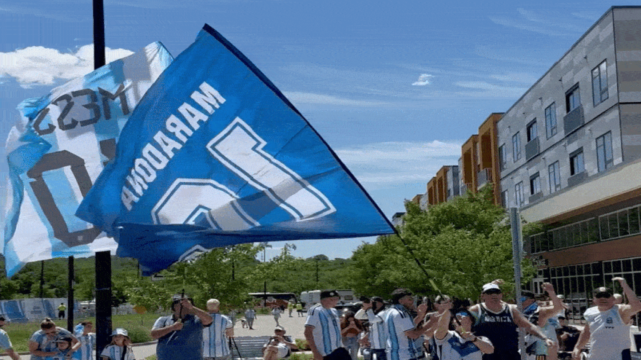 Fans in light-blue and white striped jerseys cheer on a city street, waving a large blue pennant with white numbers; modern buildings and green trees in the background.