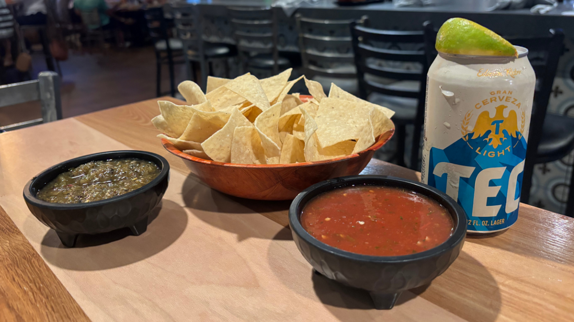 Chips and dips on a wooden table: a bowl of tortilla chips, a red salsa, and a green chunky dip; a Tecate Light beer can with a lime wedge sits nearby as a busy bar background hums.