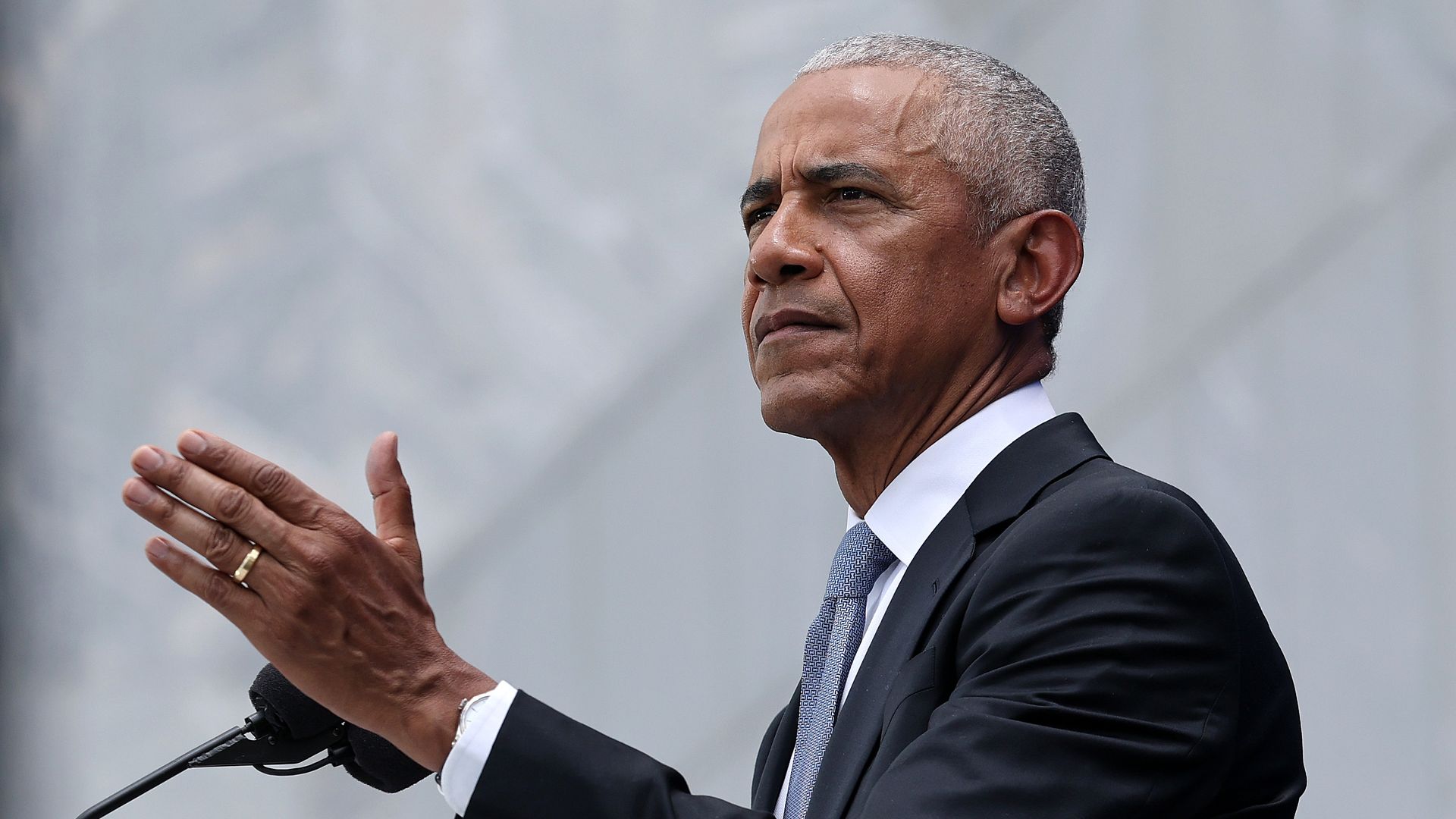 A middle-aged man in a dark suit and light blue patterned tie stands at a podium, gesturing with his hands in front of a light gray backdrop.