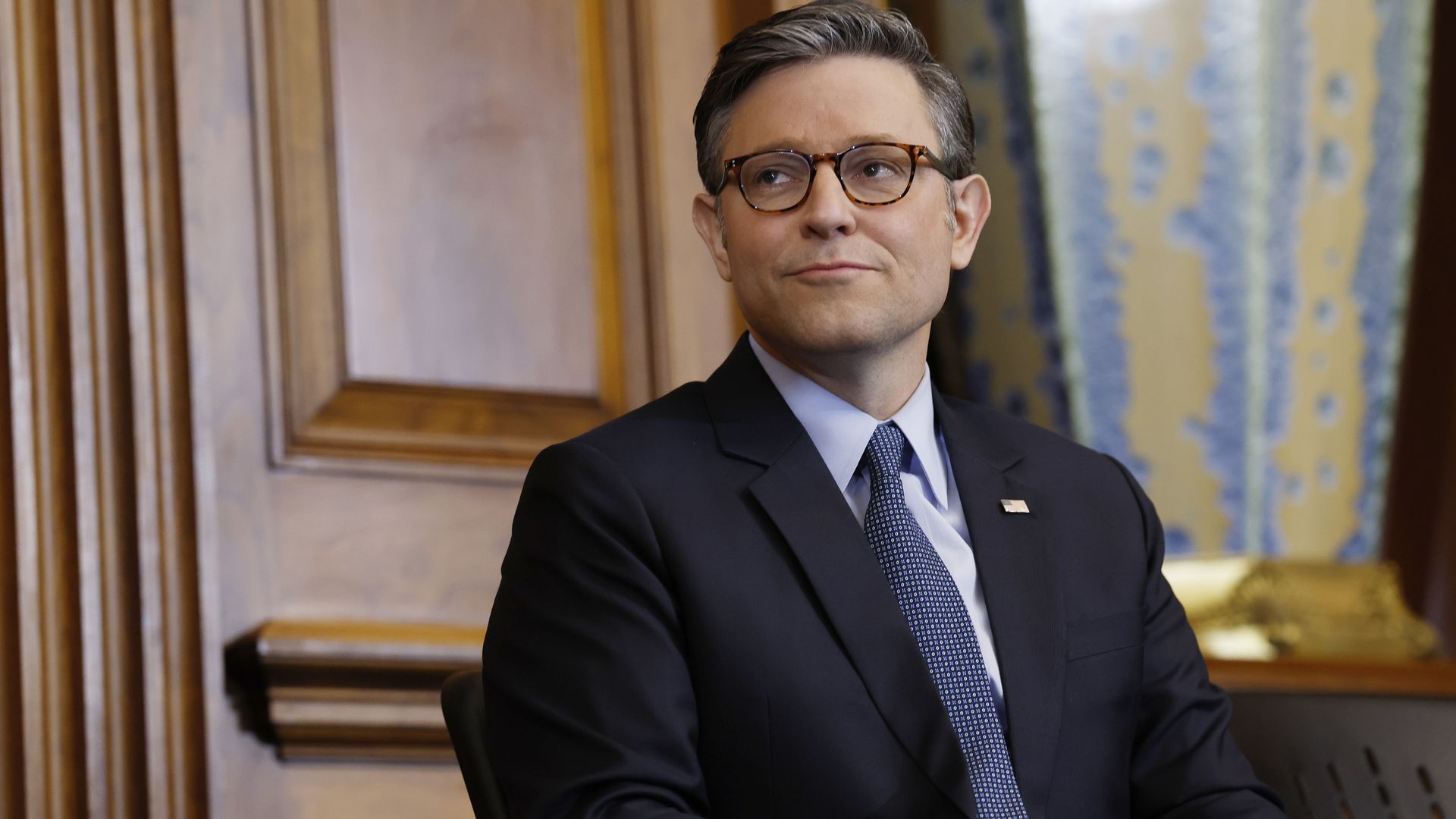 House Speaker Mike Johnson (R-La.), wearing a gray suit and sitting in a chair in front of a wooden wall.