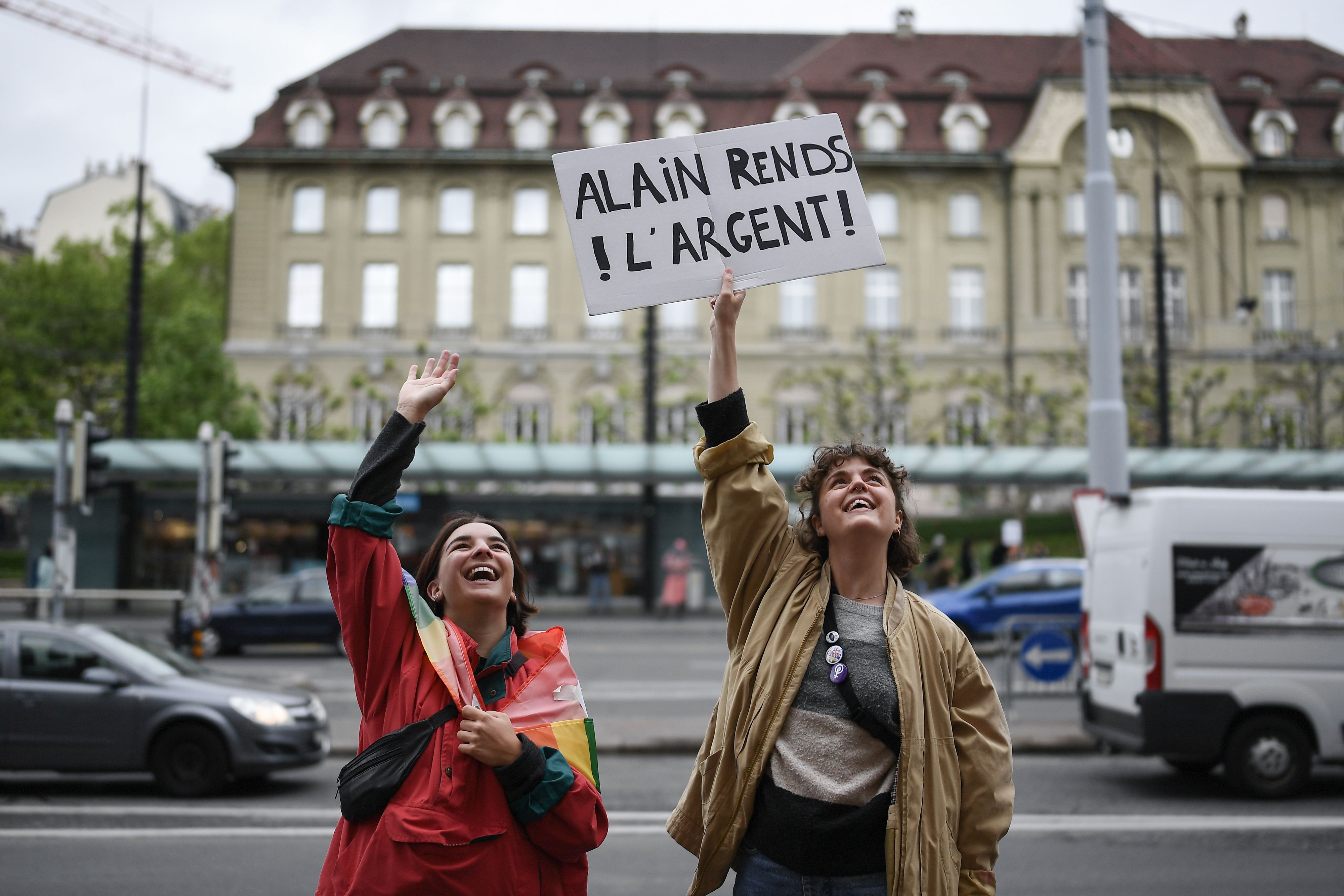 In this image, two women stand outside, one holding a sign that reads" Alain Rends L'argent!" 