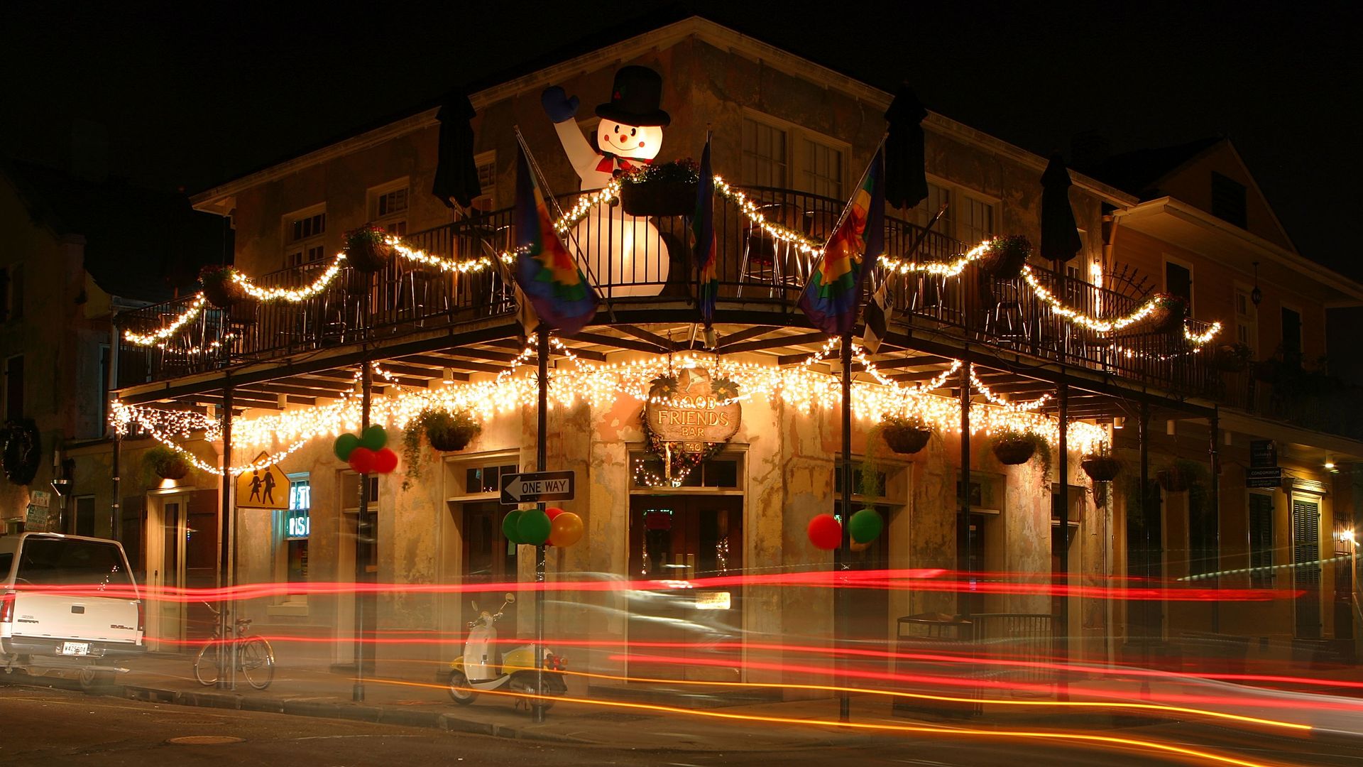 A French Quarter corner bar is photographed at night. Christmas lights decorate the balcony, and the red tail lights of a car are visible as it passes by.