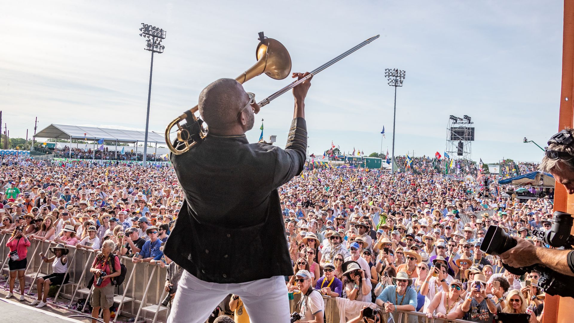 Trombone Shorty performs at Jazz Fest. He is viewed from behind so the festival crowd, standing shoulder to shoulder for as far as the viewer can see, takes up most of the background.
