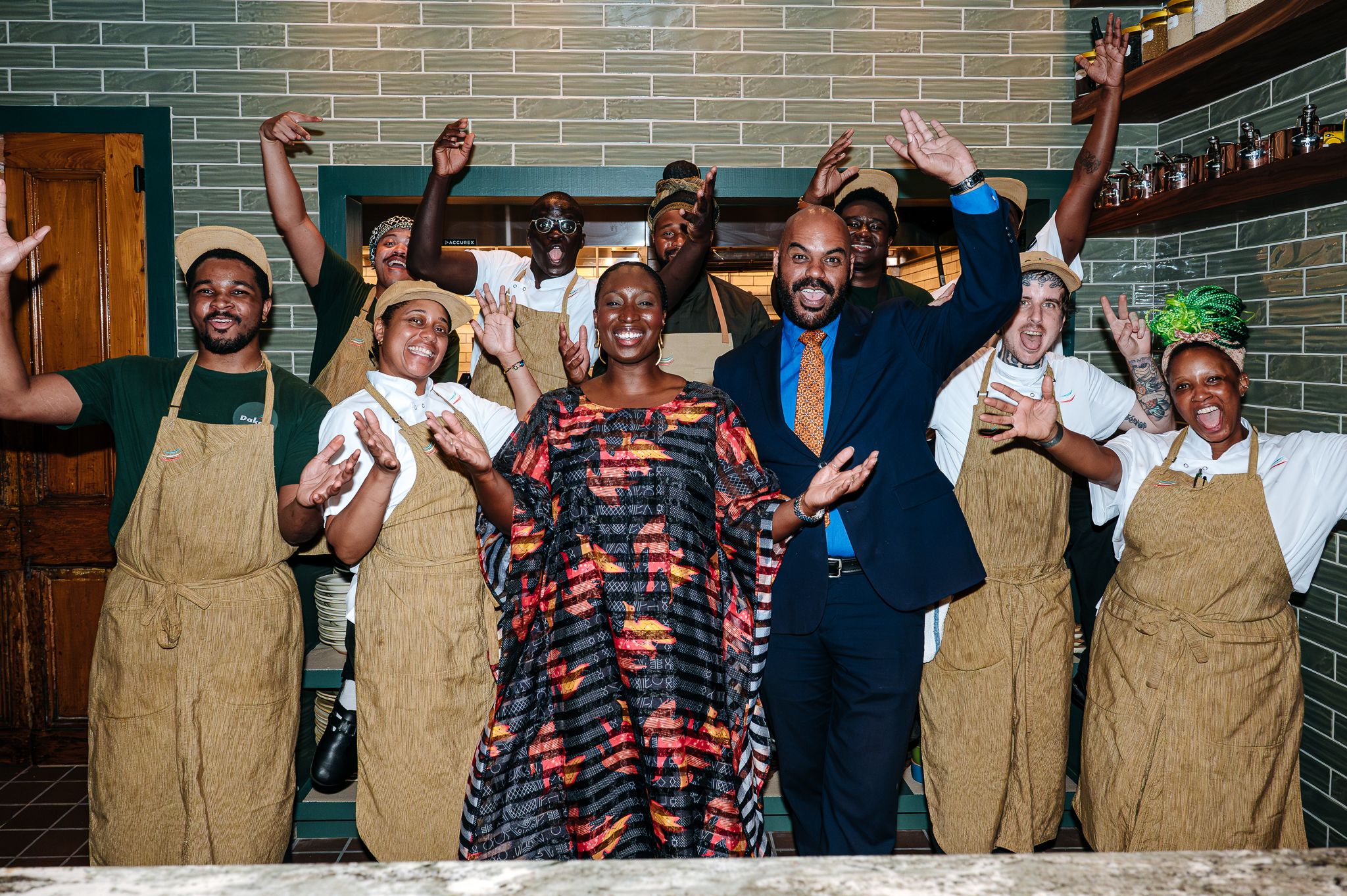 Group of people in aprons cheer in a kitchen/bar, arms raised. Front center woman wears a colorful dress; a man in a blue suit and orange tie smiles beside her, tiled walls in the background.