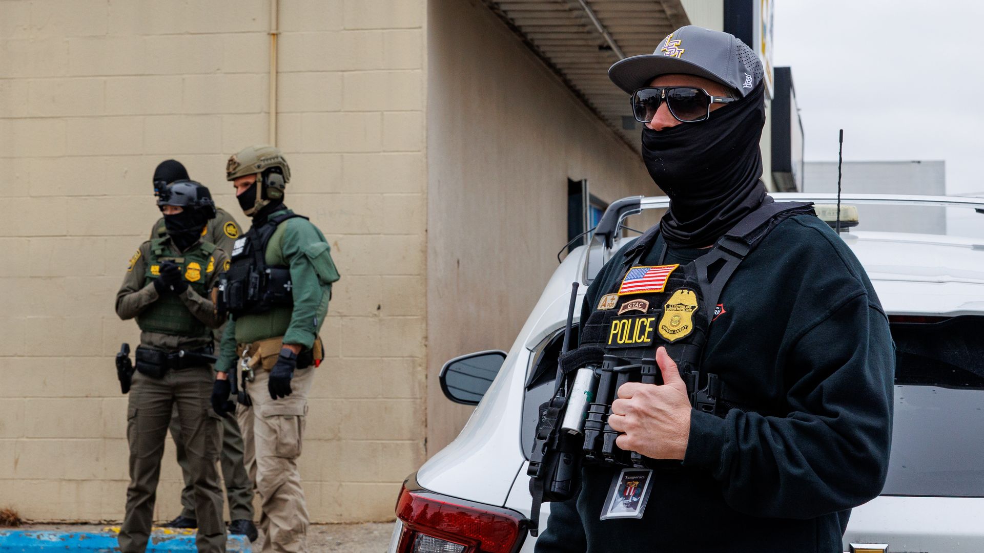 Three law enforcement officers in tactical gear stand near a white Chevrolet police vehicle outside a beige building on a cloudy day.