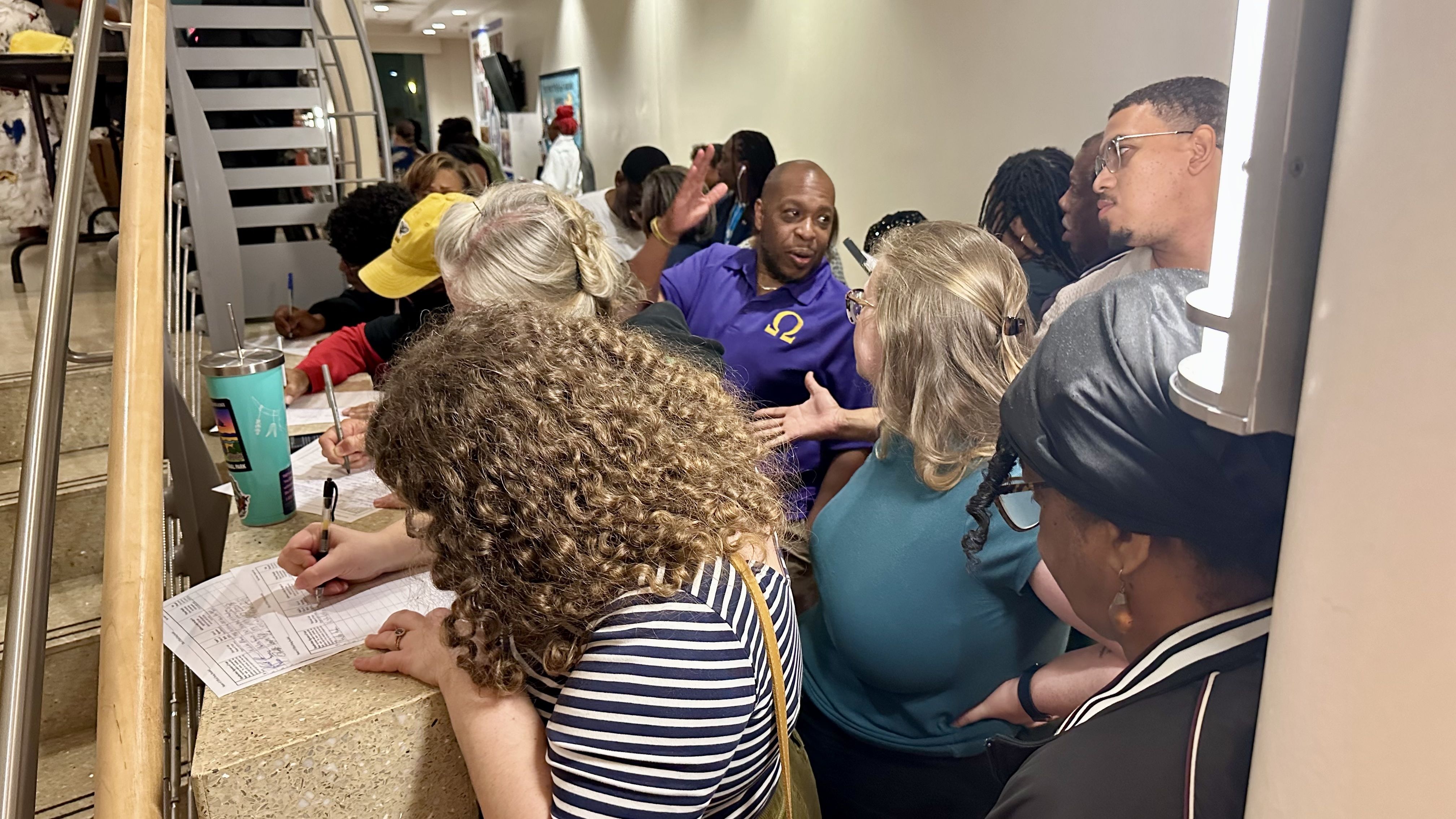 People sign papers  in a bright hallway. A man in a purple Omega shirt chats with attendees; foreground shows curly hair, a striped shirt, and a teal tumbler with a straw.