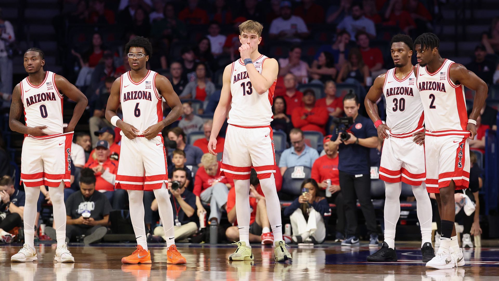 Five basketball players in white University of Arizona uniforms with red trimming stand on a basketball court during a game. 