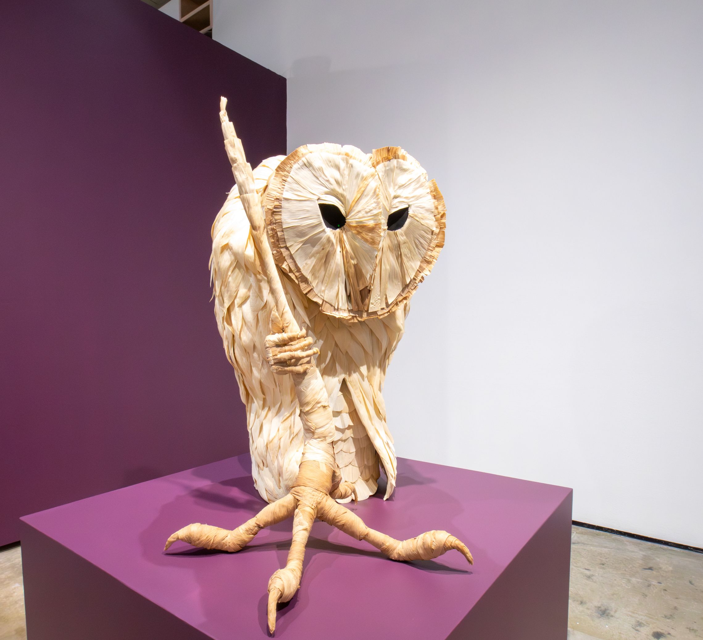 Sculpture of a stylized owl made from light beige woven materials, standing on a purple platform with a purple and white background.