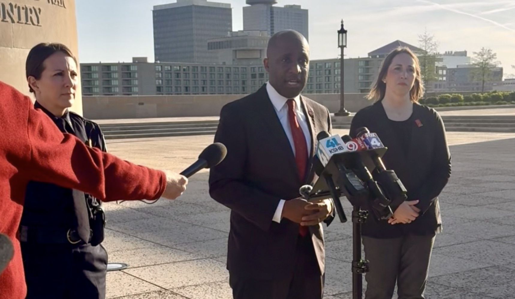 A man in a dark suit and red tie speaks at a press briefing outdoors, flanked by two women; microphones sit on a stand in front of him, with a city plaza and modern buildings in the background.