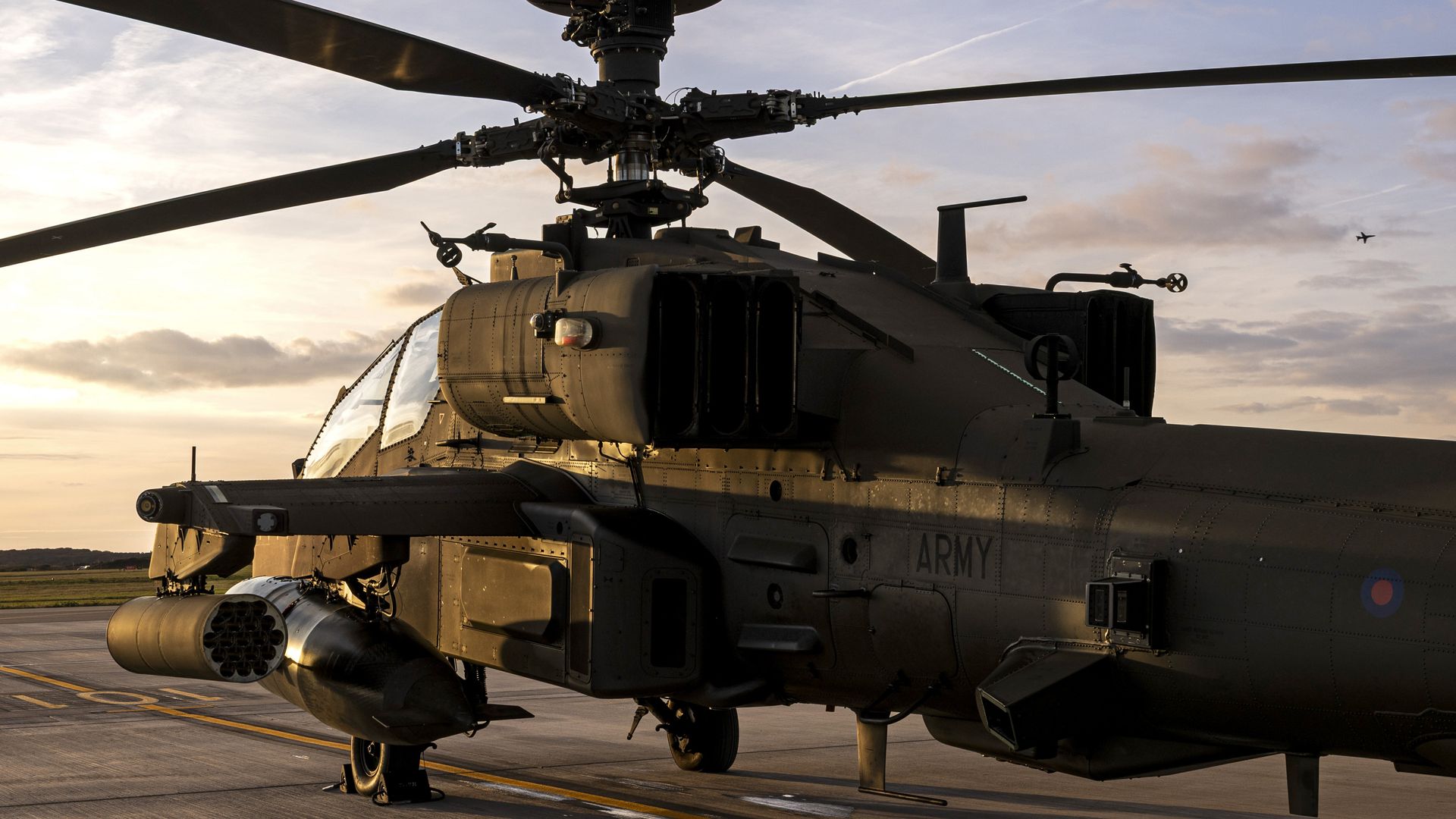 Dark military attack helicopter with rotor blades extended, parked on runway during sunset, showing detailed body with weapons and "ARMY" marking on side.