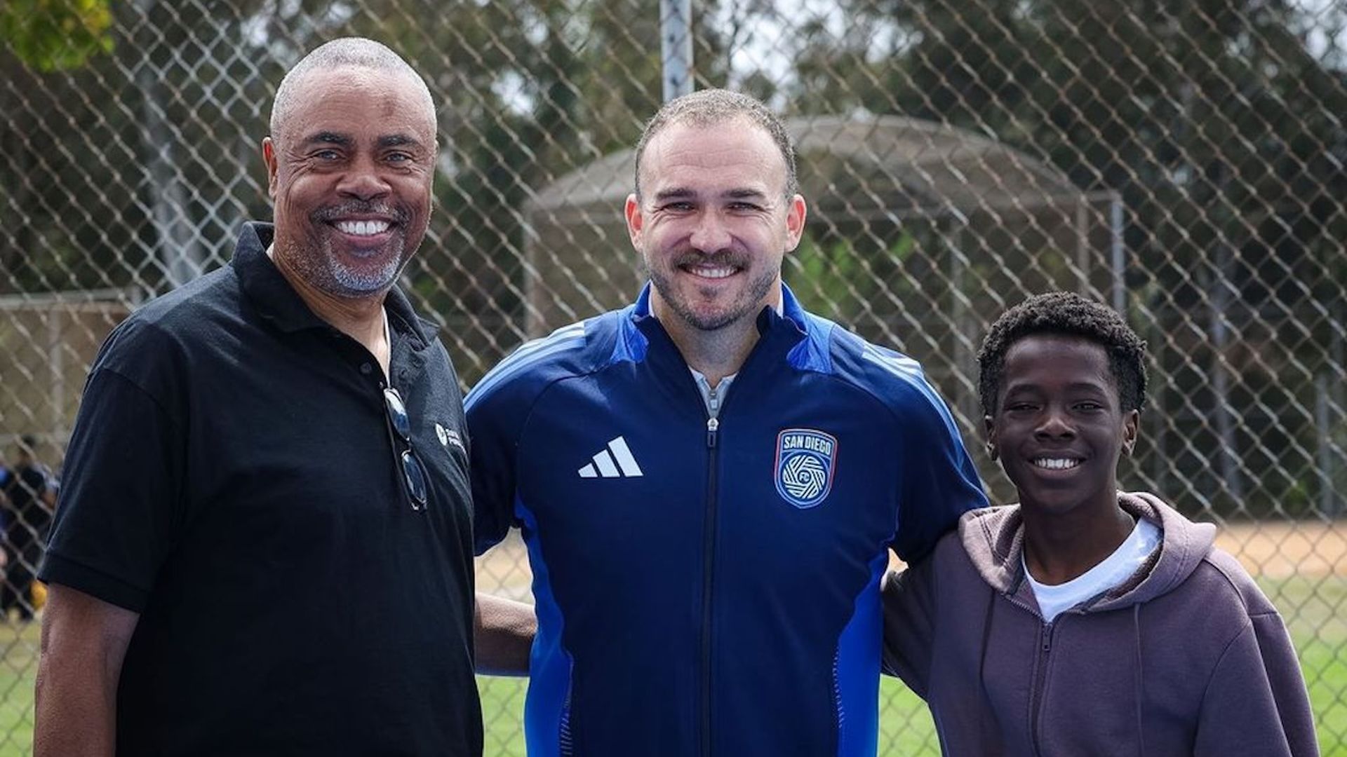 Three smiling people stand with arms around each other in front of a chain-link fence on a sports field; left man in black polo, middle man in blue jacket with a crest, right boy in a purple hoodie.