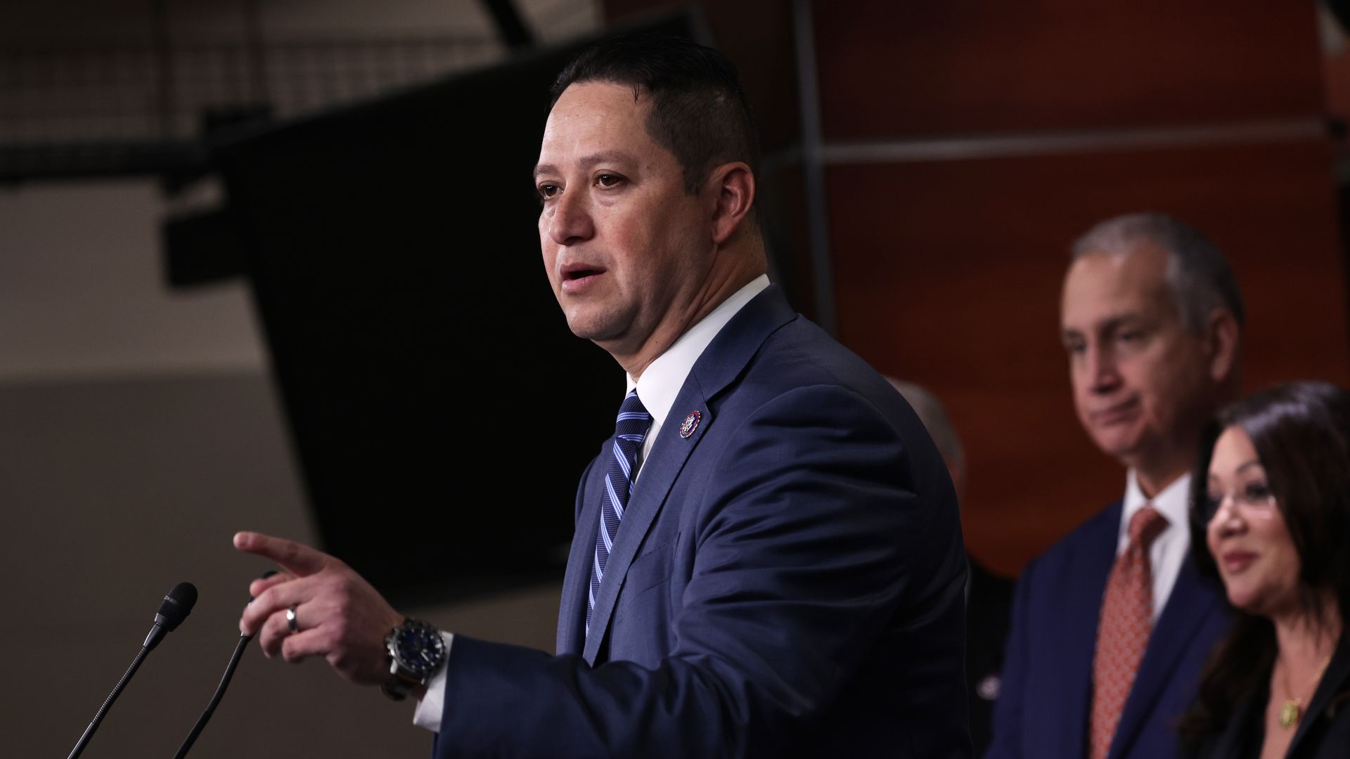 Man in a navy suit and striped tie speaking and gesturing at a microphone, with two people standing behind him against a dark and wood-paneled background.