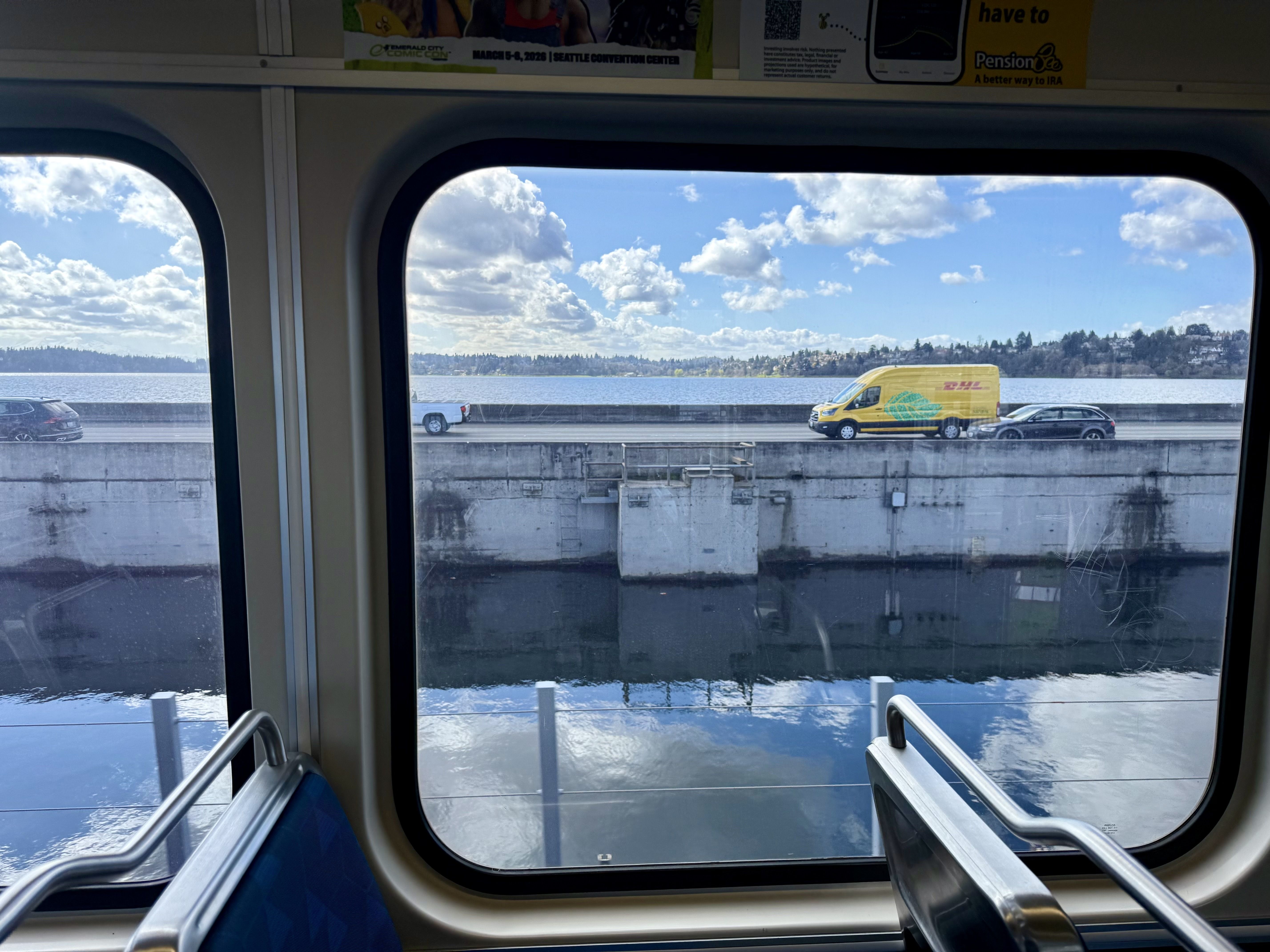 Inside a train carriage, looking through a large window at a sunny waterway. A yellow DHL delivery van drives on a concrete road beside the water, under a blue sky with white clouds.