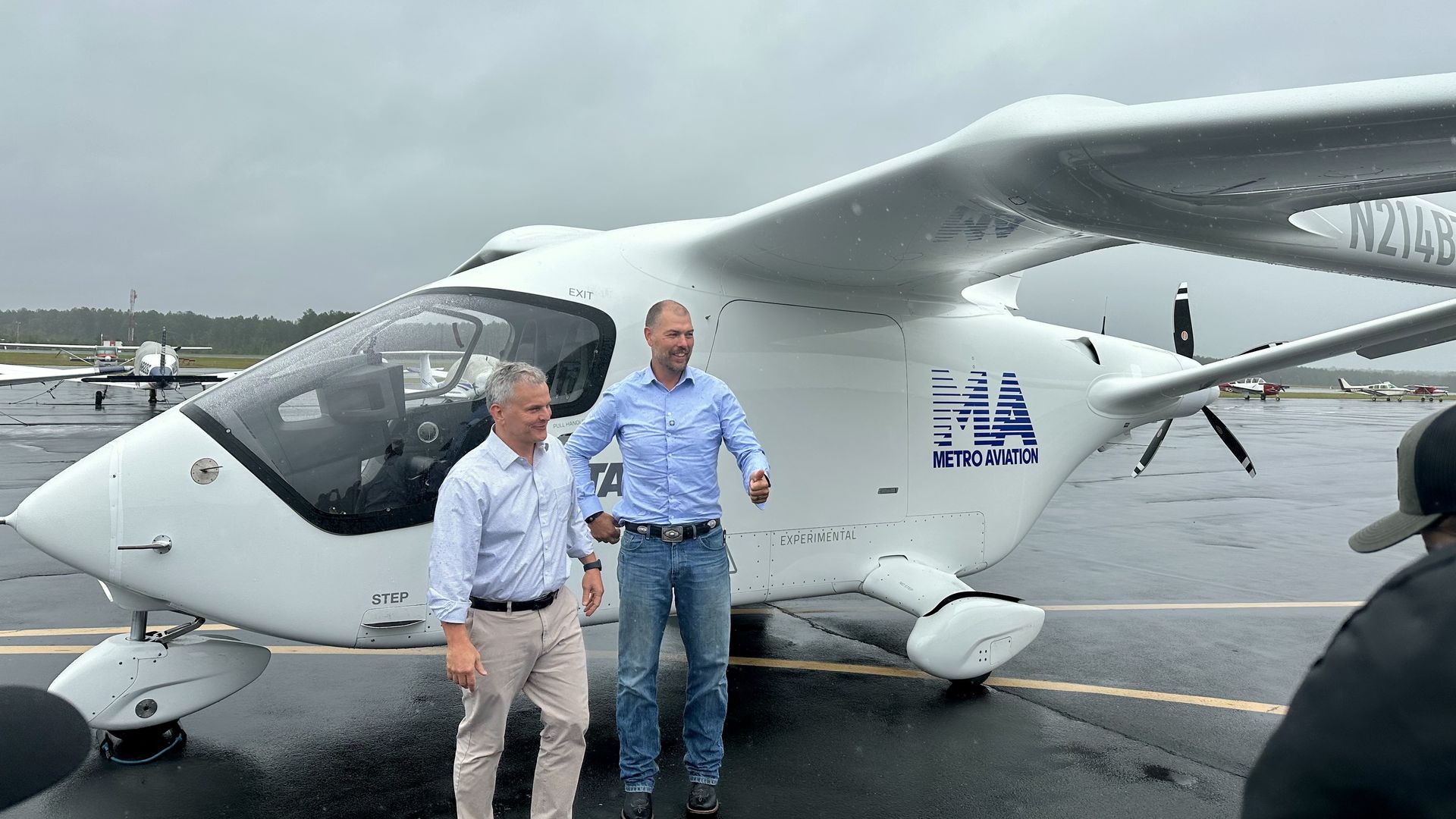 Two men stand beside a white Metro Aviation experimental helicopter on a wet tarmac under a cloudy sky; cockpit, nose, and tail are visible with distant small aircraft.
