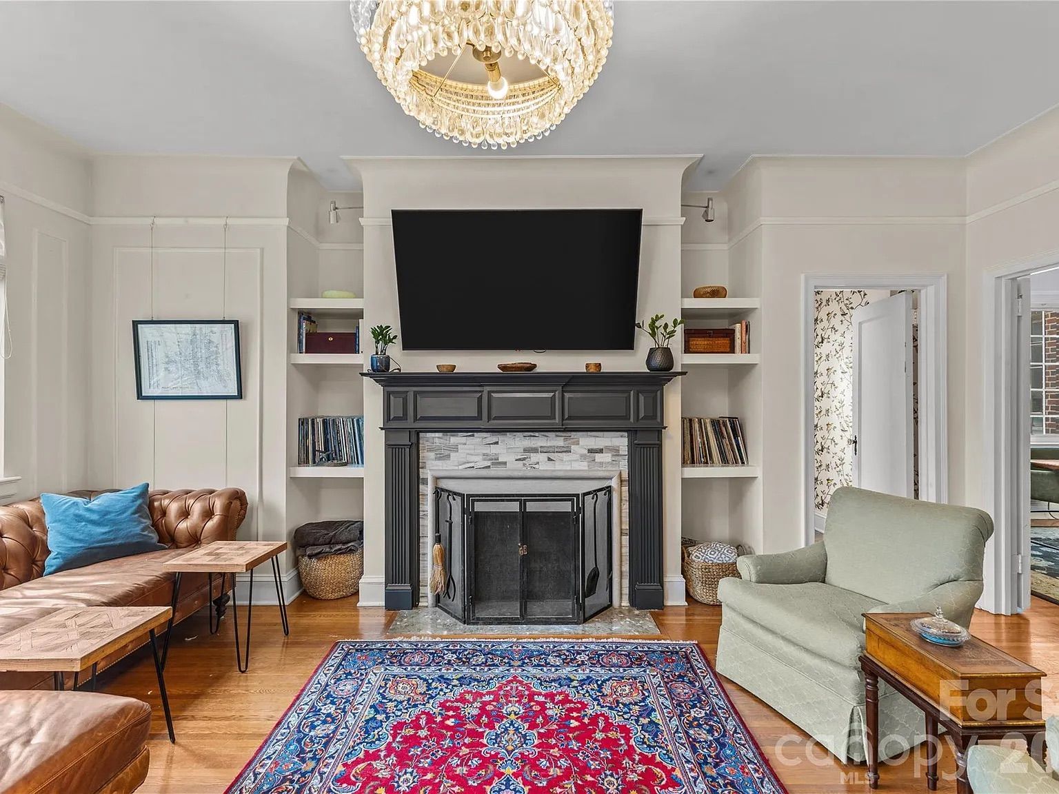 Living room with white walls, a black fireplace, a mounted TV, brown leather sofa with a blue pillow, a green armchair, patterned red and blue rug, wooden tables, and a chandelier.