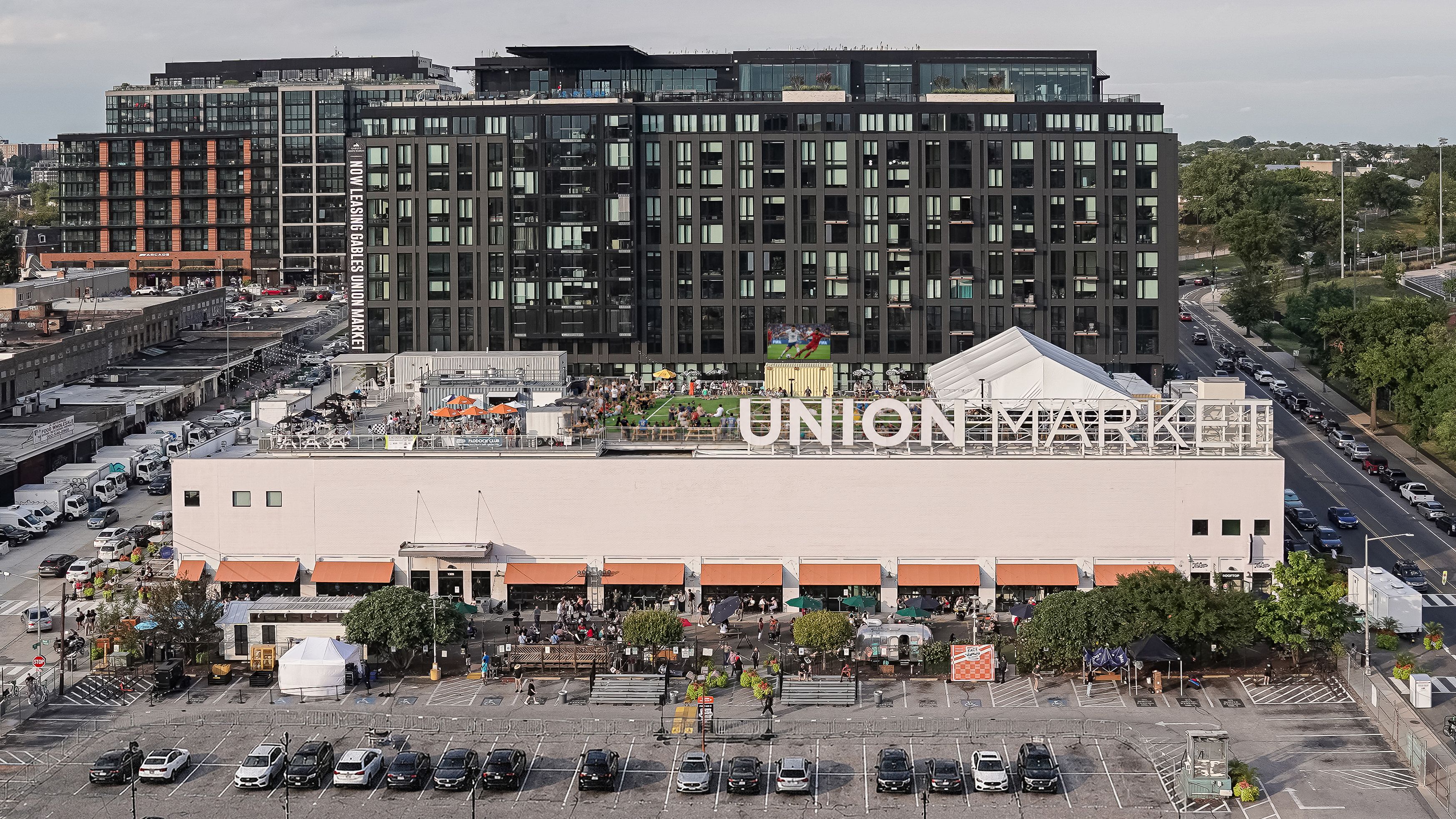 An aerial view of a modern dark glass Union Market building with a white base, an outdoor event area with orange umbrellas and tents, and a vertical banner advertising leases.