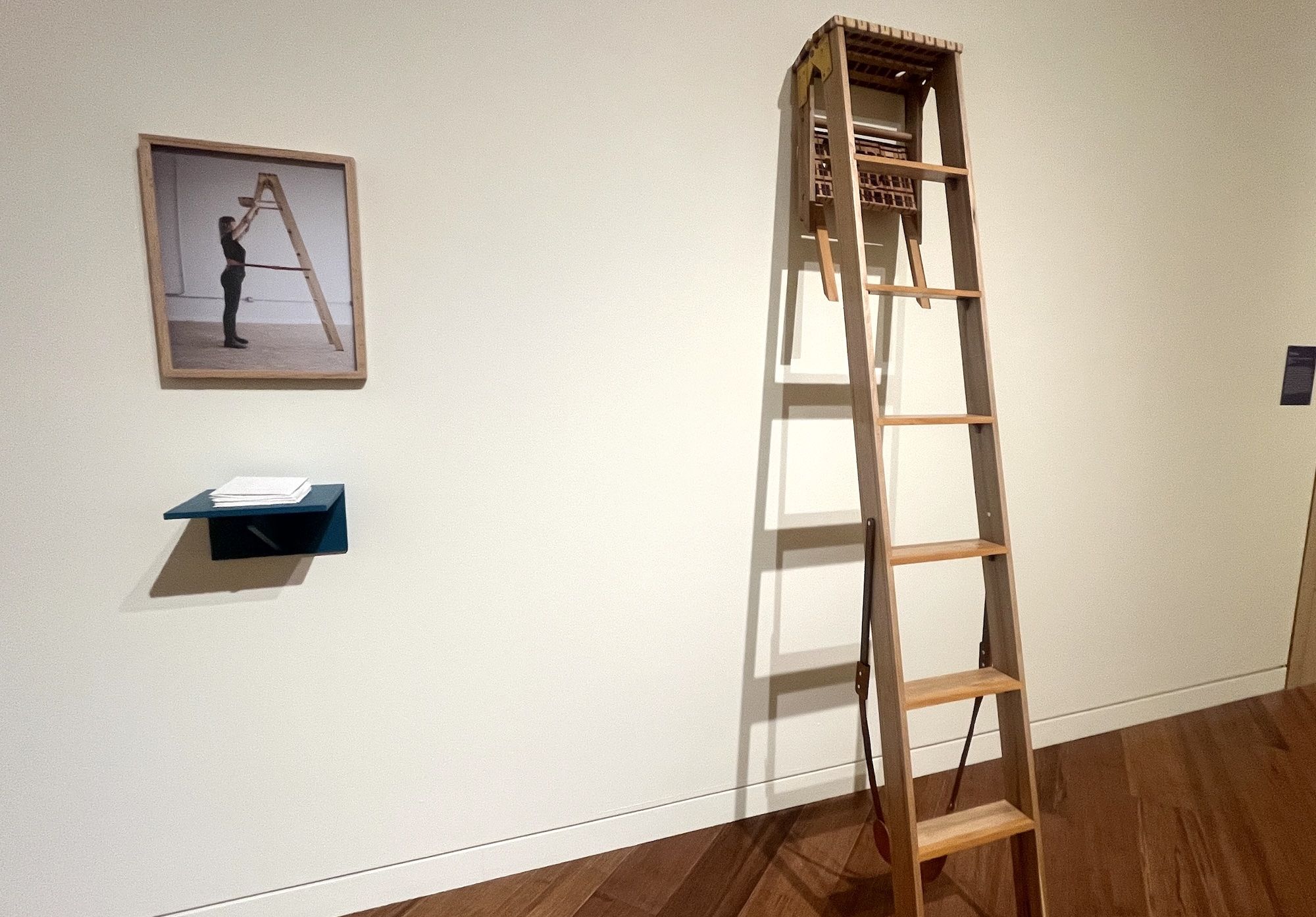 Wooden ladder mounted unusually high on a white wall in an art gallery, with a framed photo of Crist interacting with the ladder and a small shelf holding folded papers nearby.