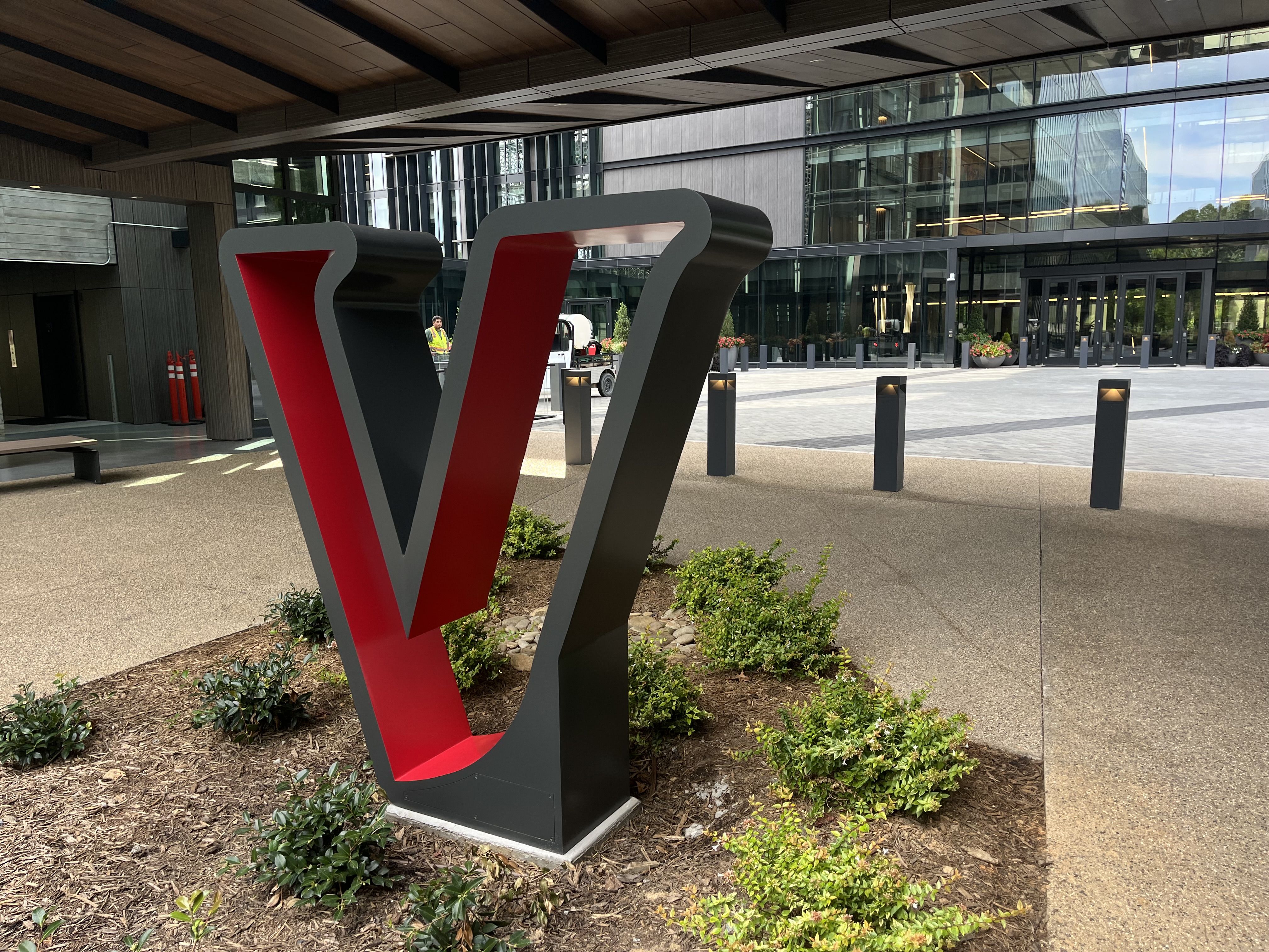 Large black and red sculpture of the letter V outdoors near a modern glass building, surrounded by small green bushes and concrete pavement.
