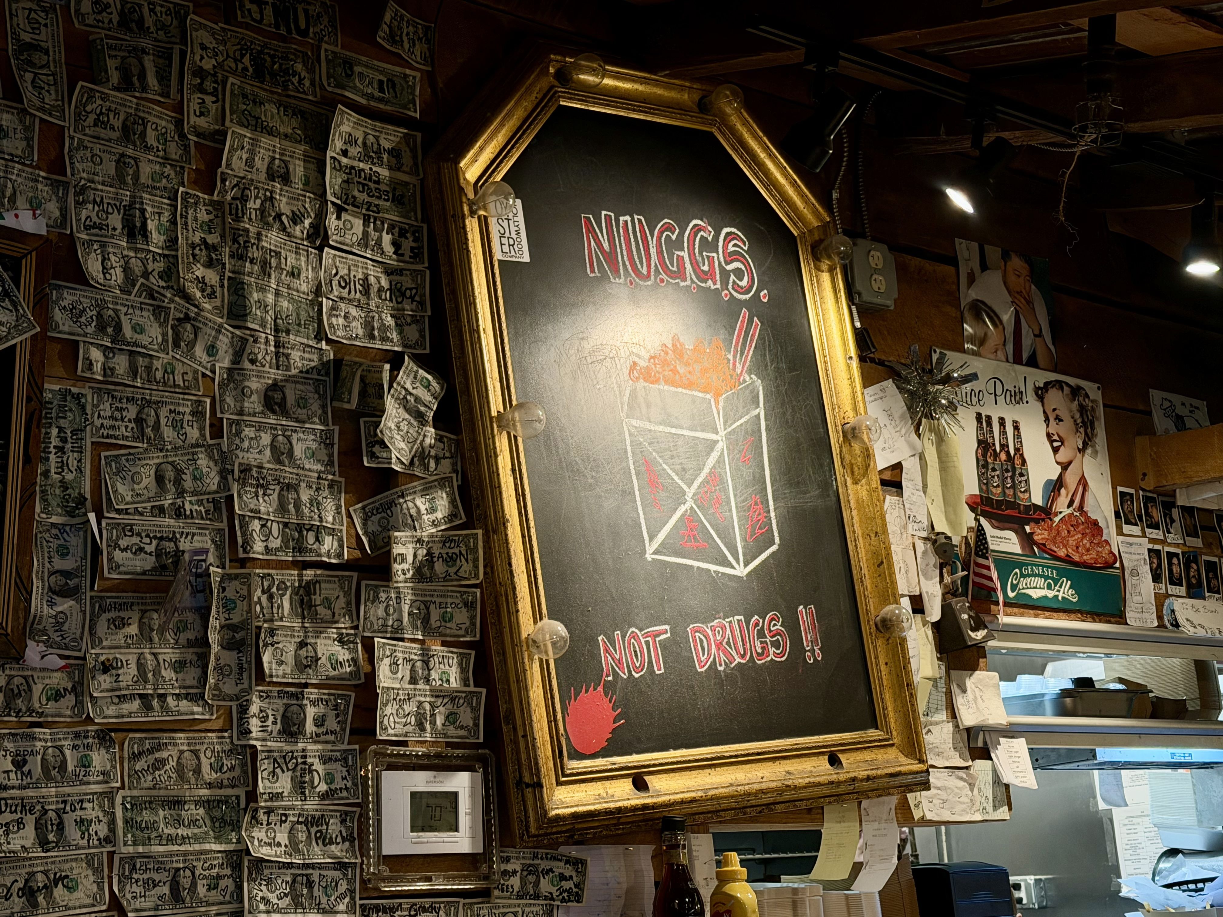 Dim bar wall with a large gold-framed chalkboard showing NUGGS in red. Left side is covered in overlapping dollar bills, right side has vintage posters; warm bulbs frame the chalkboard.