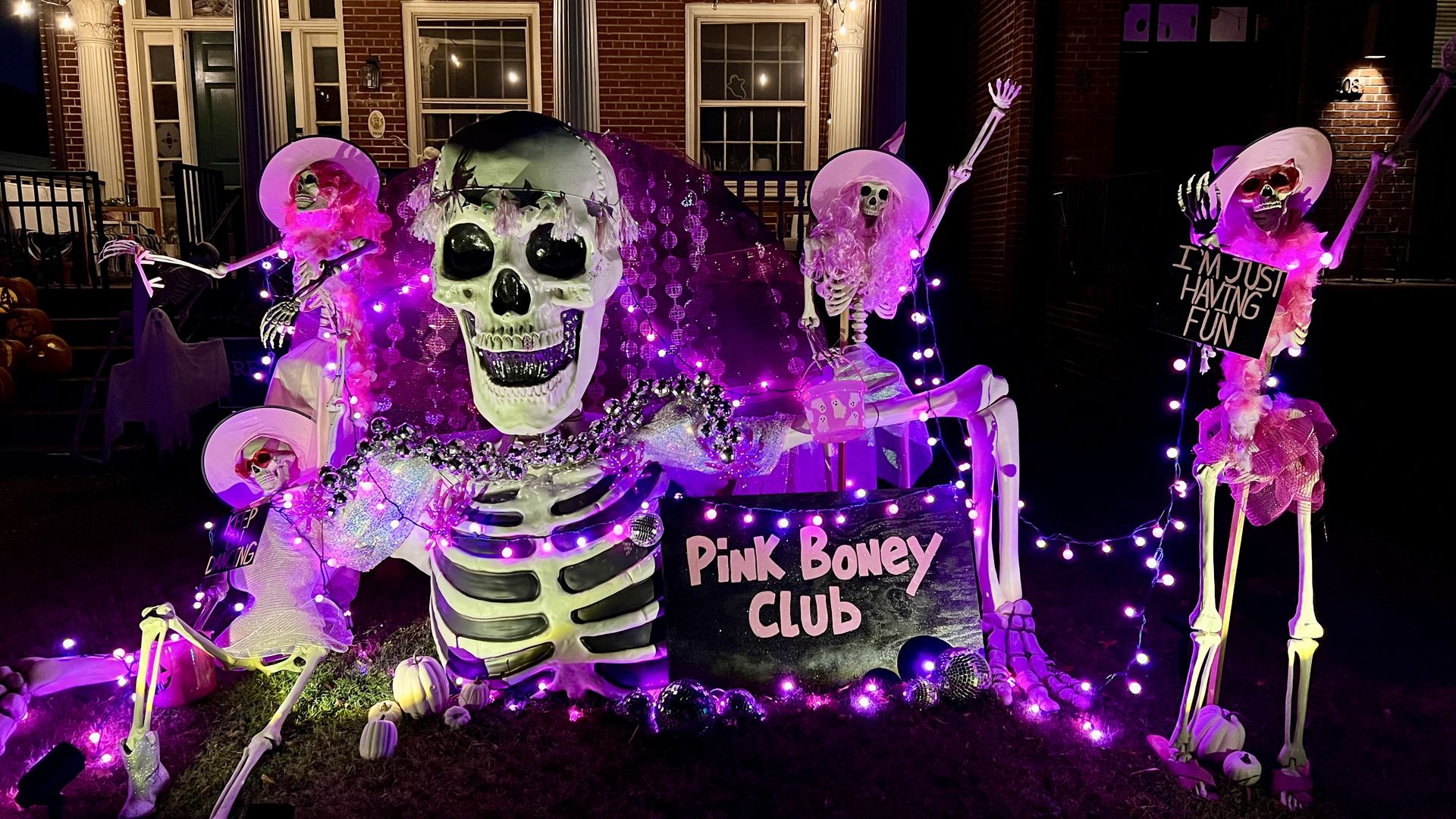 A pic of halloween decor showing a giant skeleton surrounding by dancing skeletons wearing pink boas and cowboy hats with a sign in front that says "Pink boney club"