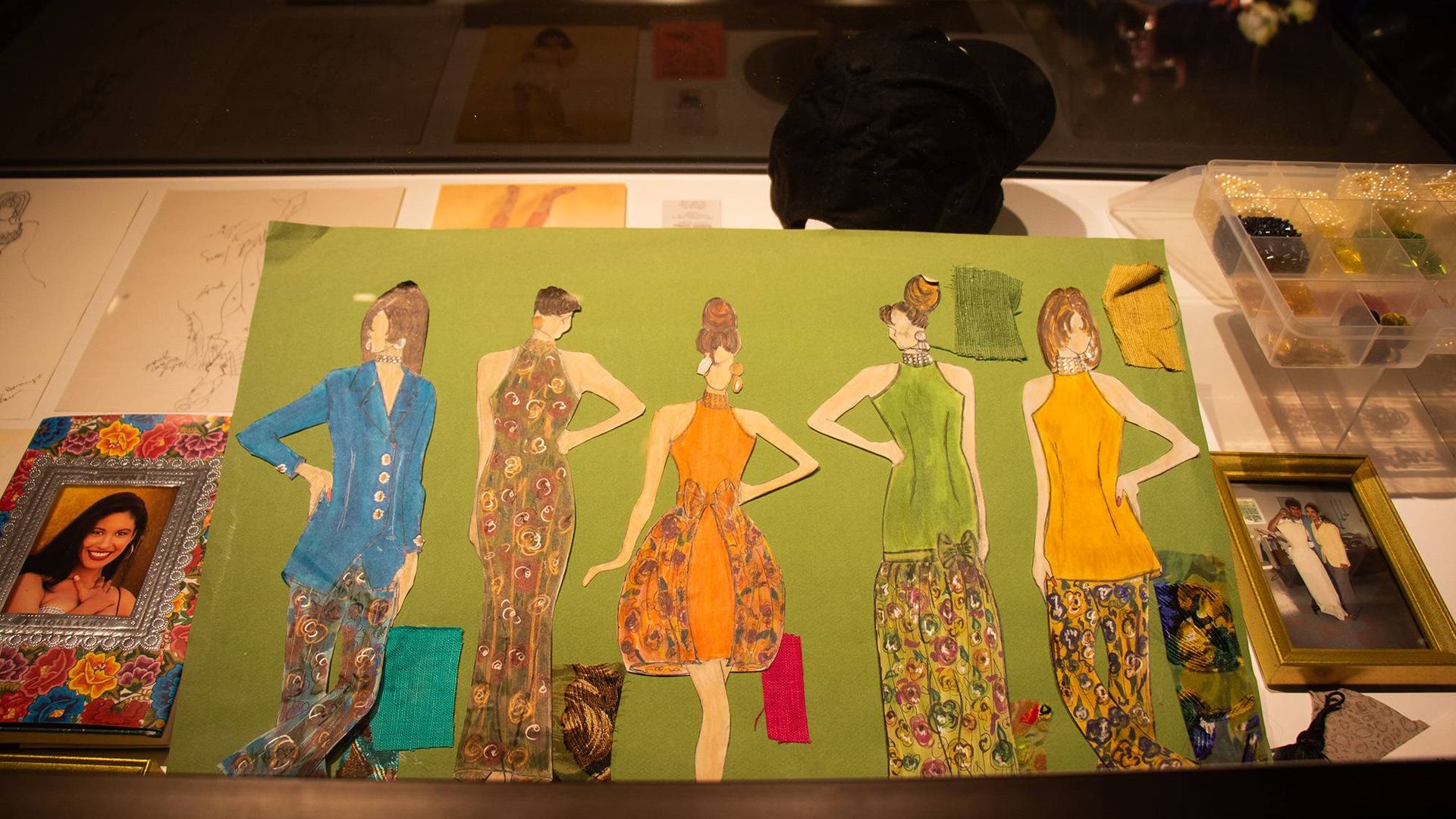 Fashion sketches of five women on green background, dressed in colorful outfits with floral patterns and solid colors like blue, orange, and green, with fabric swatches attached.