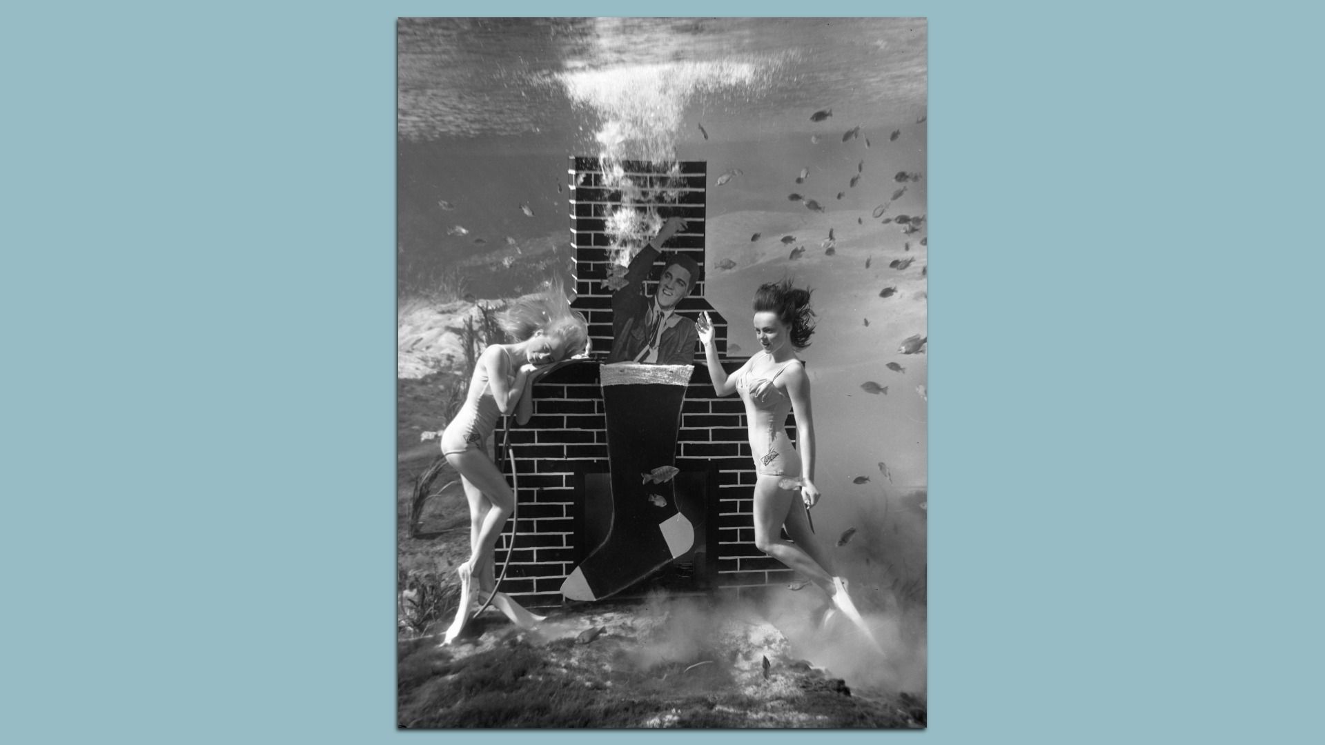 Black-and-white underwater photo of two women in swimsuits posing by a large brick fireplace prop with a cutout of a man inside, surrounded by small fish and bubbles.