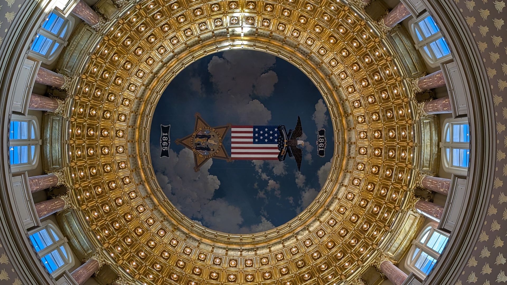 Looking up at a circular rotunda with a gold coffered dome and pink marble columns; at the center a mural shows the U.S. flag and a bald eagle against a cloudy sky, flanked by banners reading 1867.
