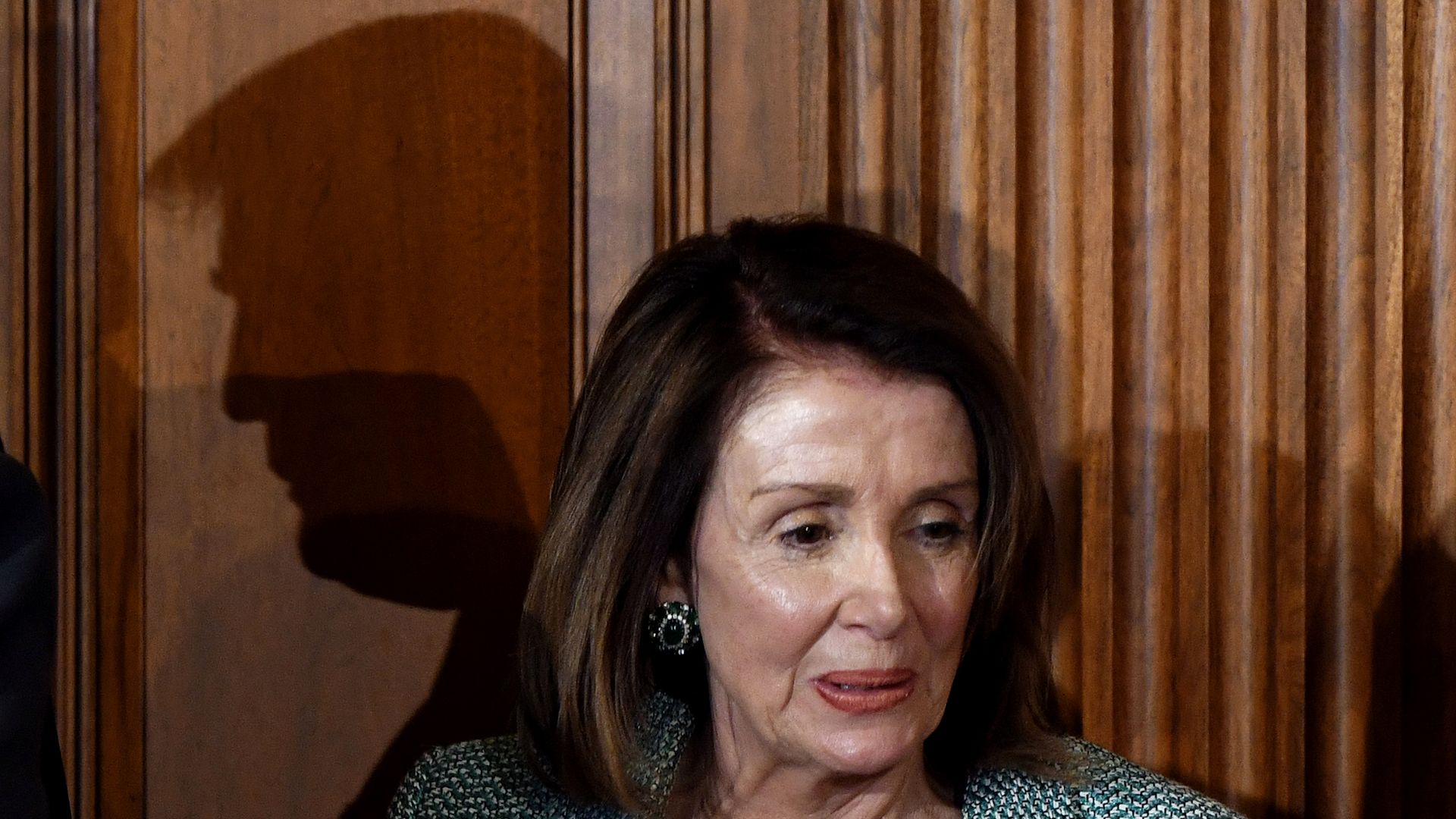 In this image, House Speaker Nancy Pelosi stands against a wooden wall with the shadow of Donald Trump behind her.