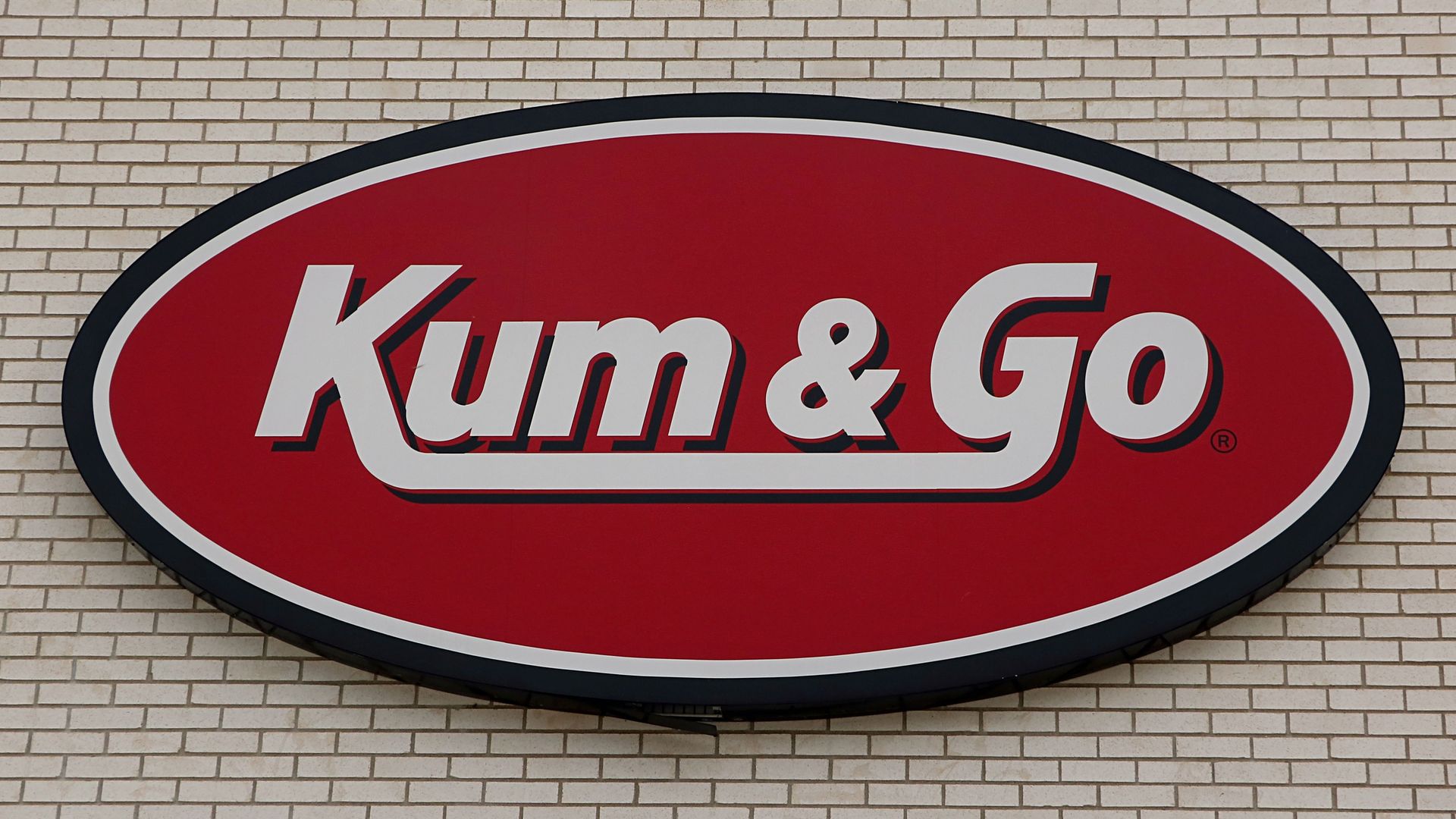 Red oval Kum & Go logo with white and black text on a light brick wall background, representing a convenience store chain.