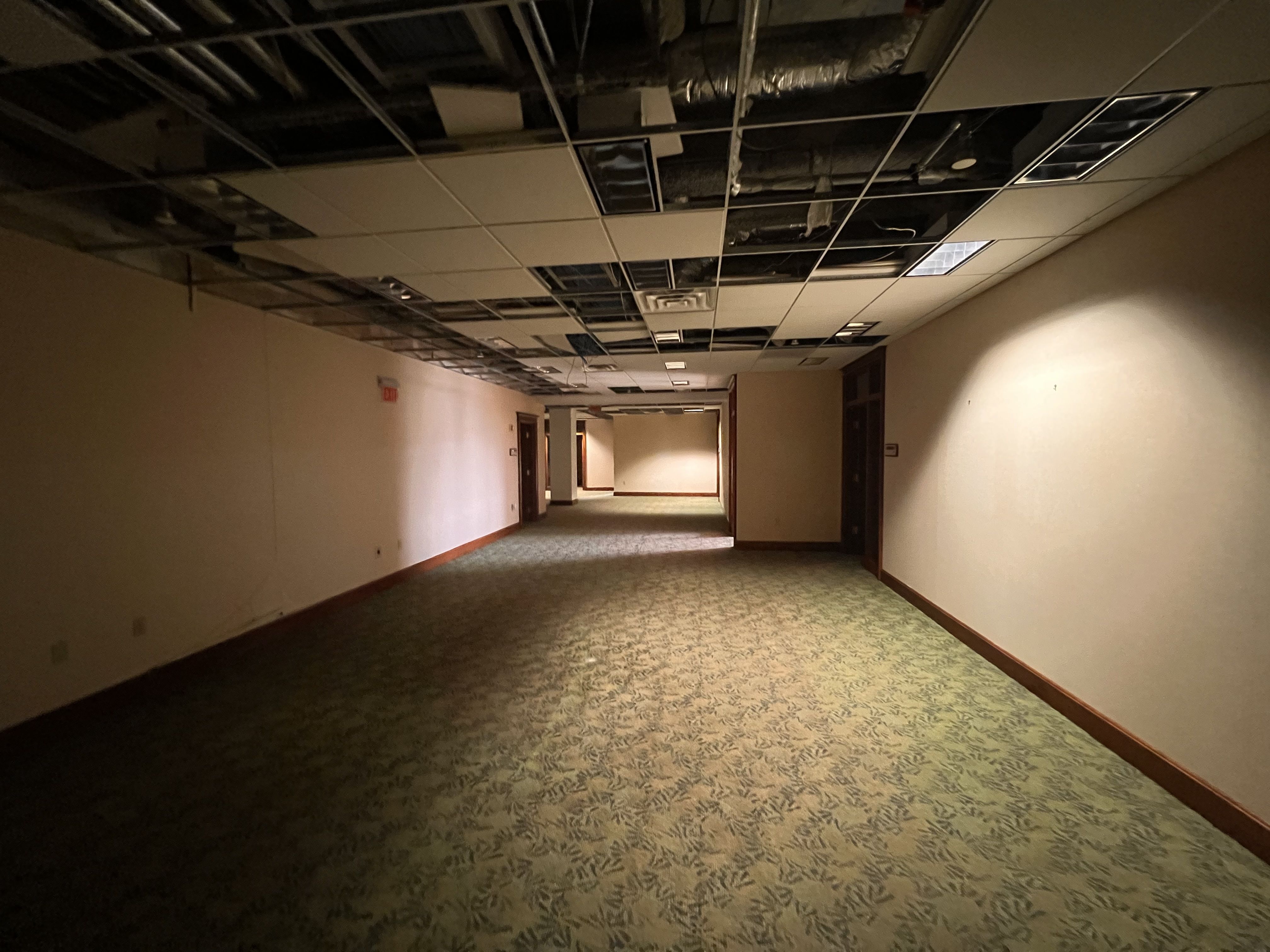 Dim hallway with a damaged drop ceiling; many tiles missing, exposing ducts and wires. Beige walls, patterned carpet, doors along both sides, and a distant exit sign.