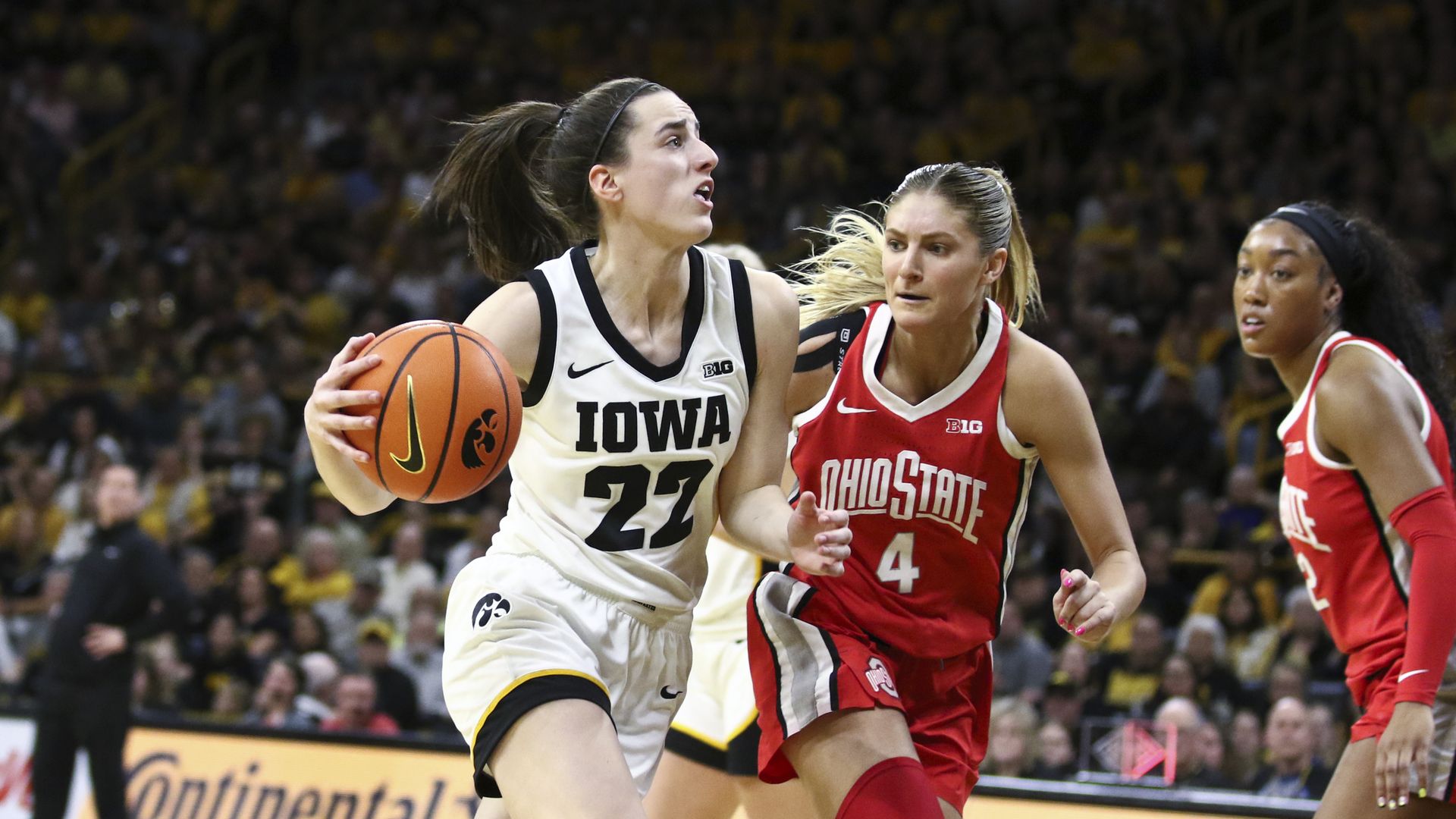 A woman in a white basketball uniform that says Iowa drives the ball against a woman in a red uniform that says Ohio State