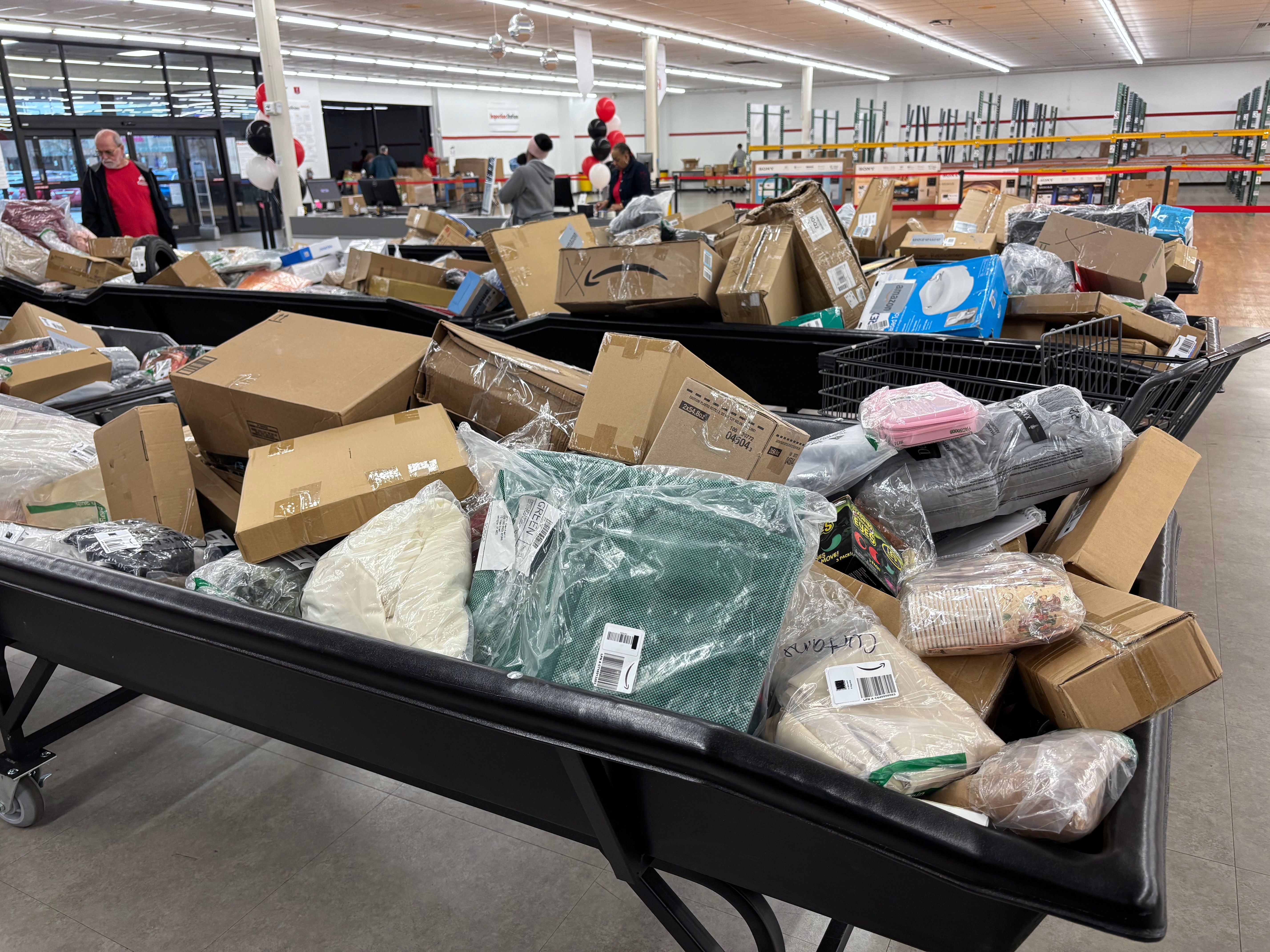 Large bins filled with assorted packed products and cardboard boxes in a spacious, well-lit store with shelves and a few people browsing in the background.