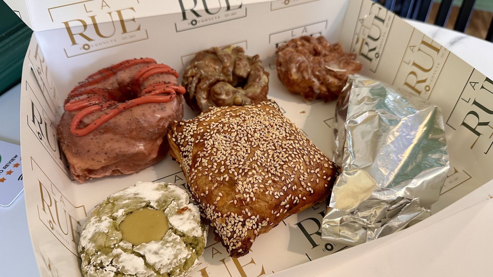 A sesame-seed bun sits foreground; behind it are crullers and a pink frosted pastry with red drizzle and foil-wrapped items in a takeout box with the words, "La Rue" printed on it. 
