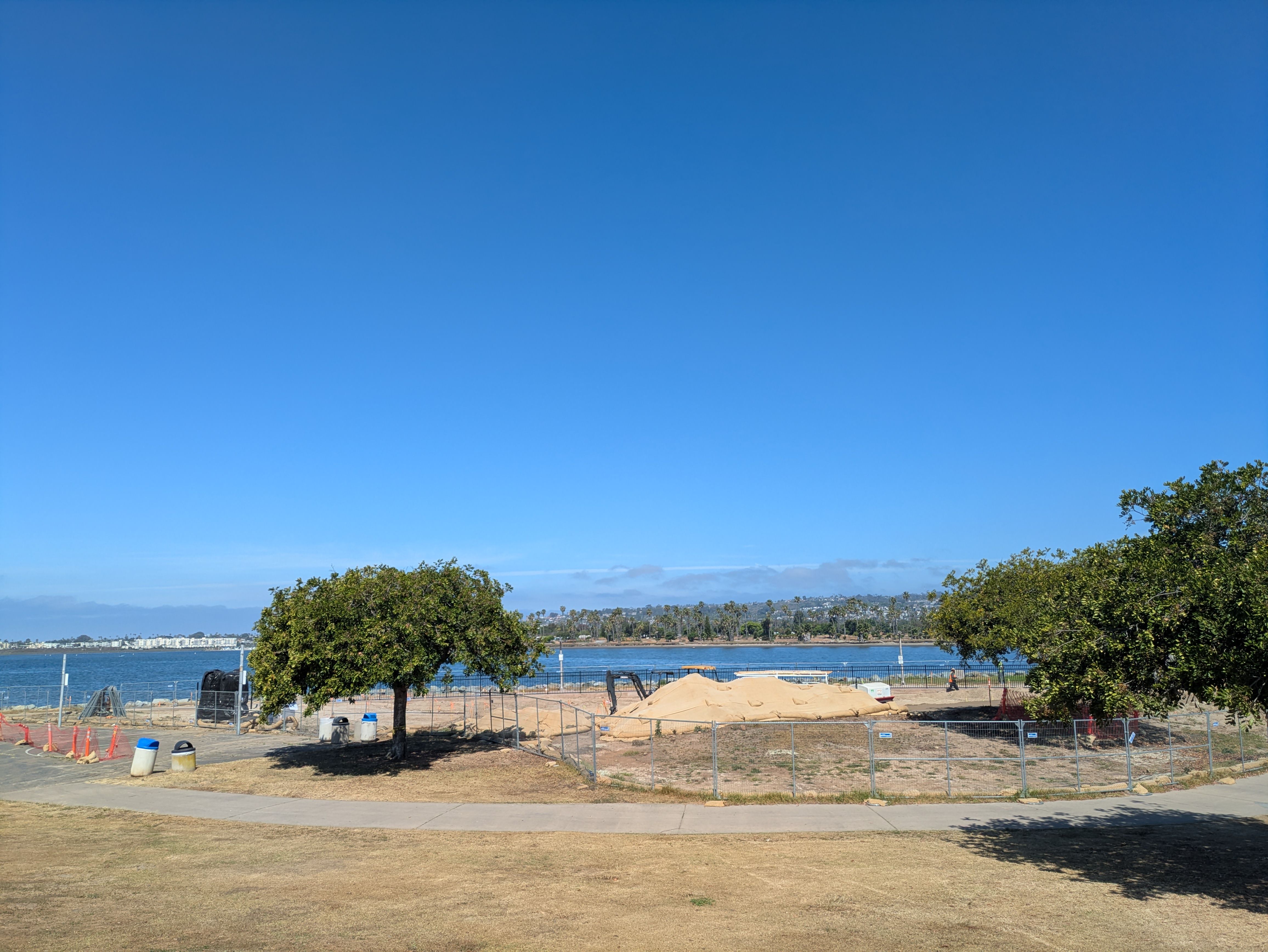 Clear blue sky over a waterfront park: two trees in the foreground, a curved path, and a fenced sandy mound near the water. Distant shoreline and buildings line the horizon.