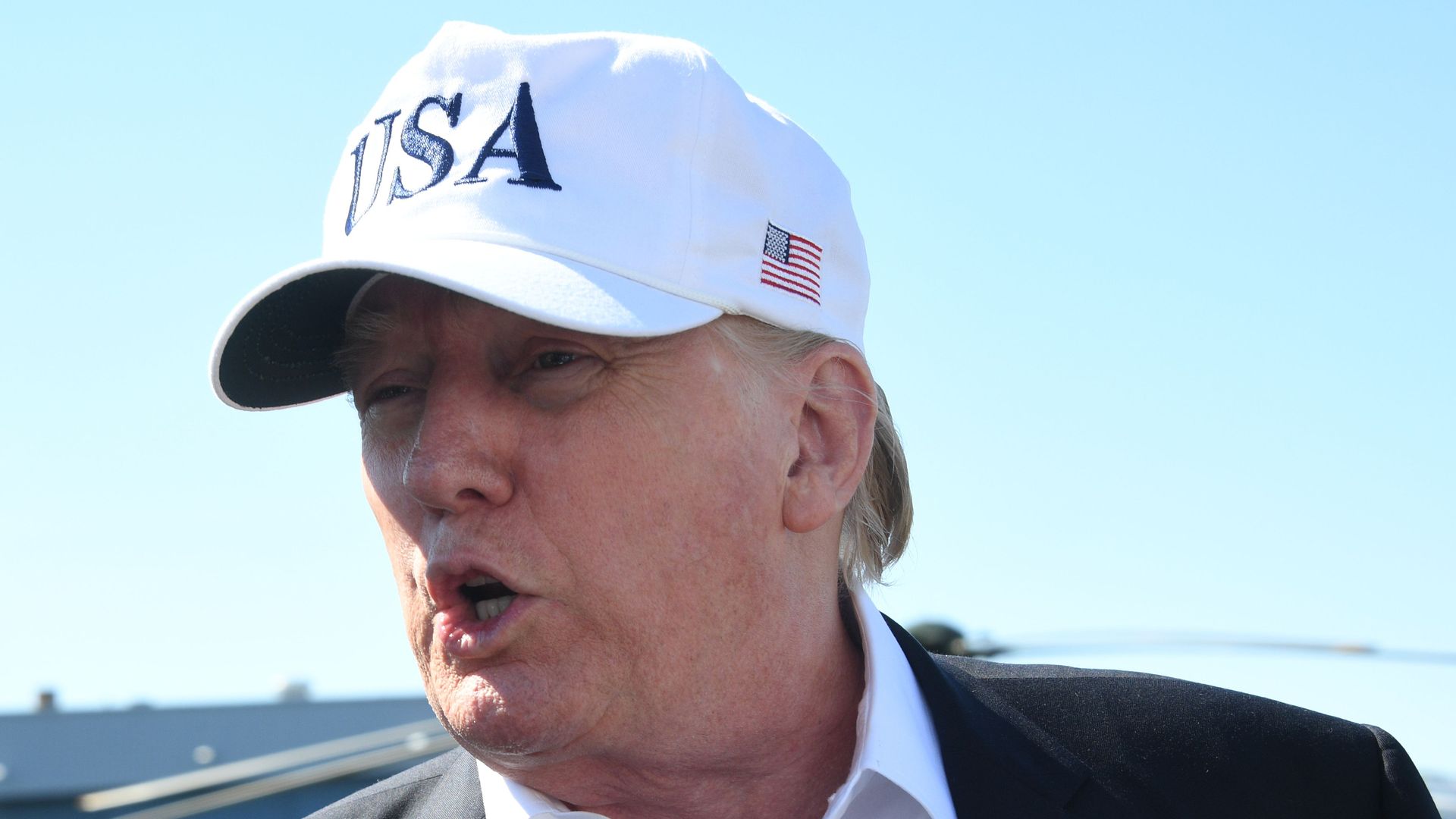 Donald Trump speaks with mouth open before a blue sky wearing a white USA hat.