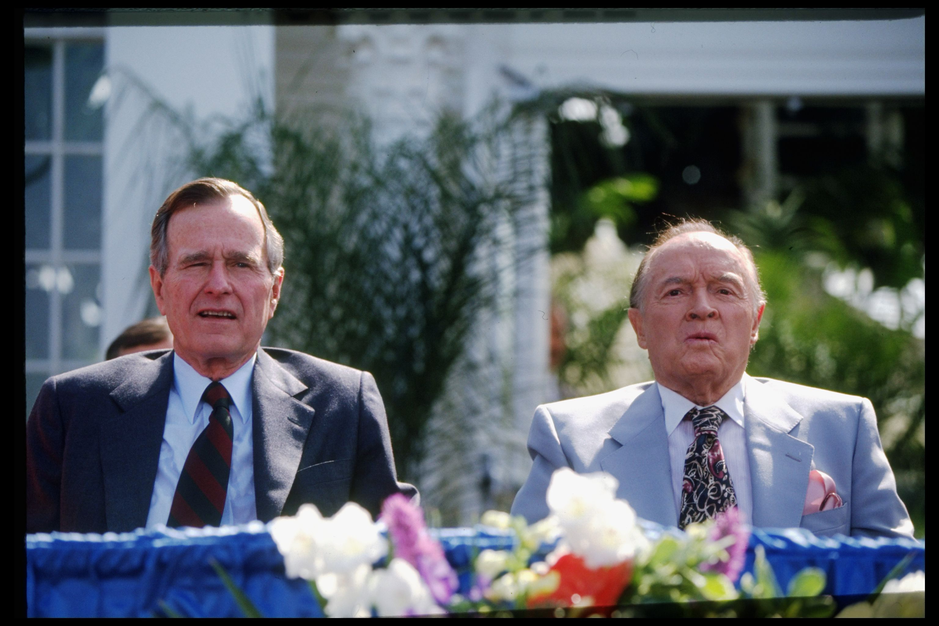 President George H.W. Bush and comedian Bob hope sit in front of a flower arrangement. 