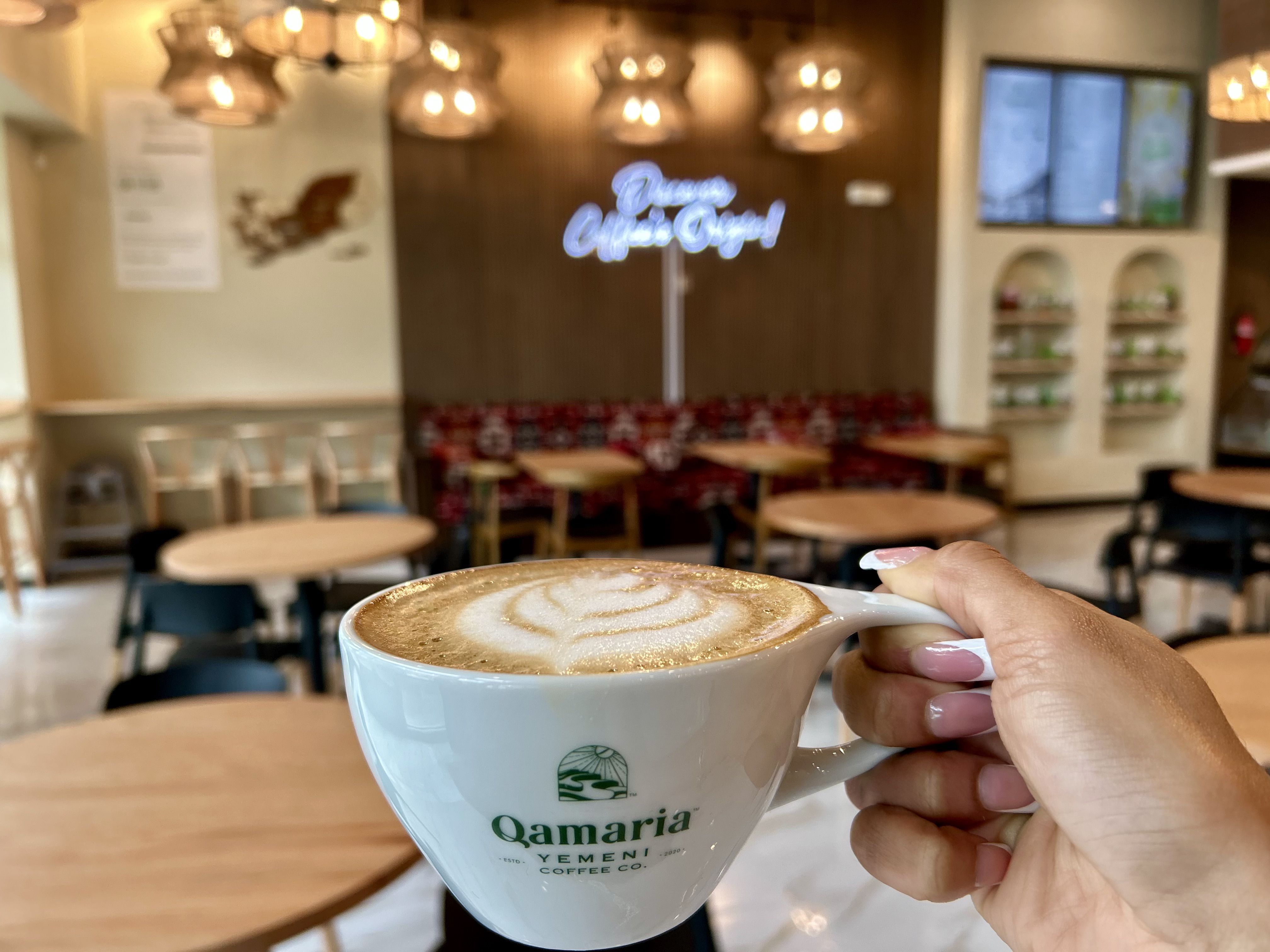 Hand holding a white cup of coffee with foam art inside a cozy cafe with wooden tables, black chairs, warm hanging lights, and a neon sign on a brown wall.