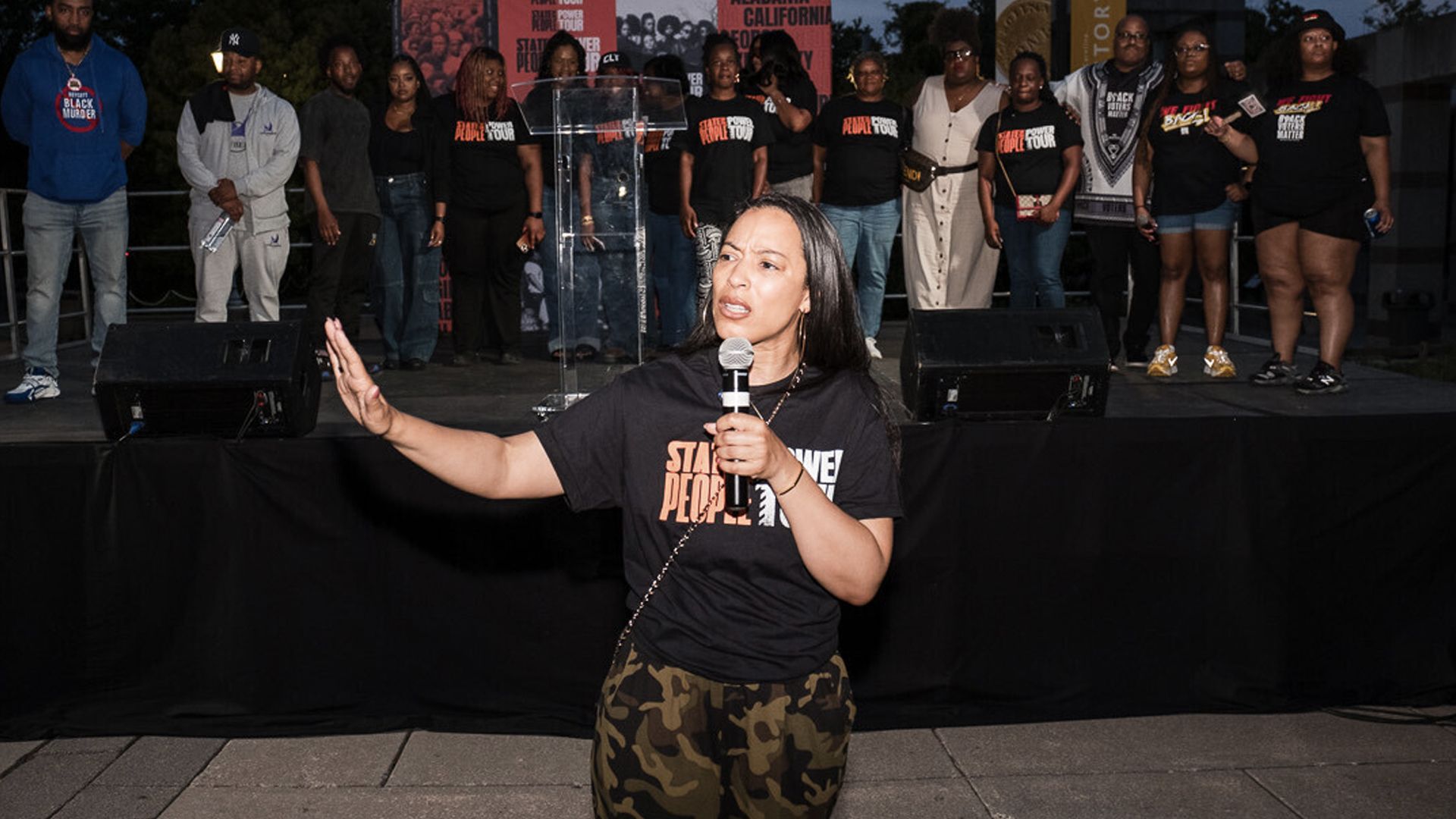 angela rye talking into a microphone while people stand behind her on a stage