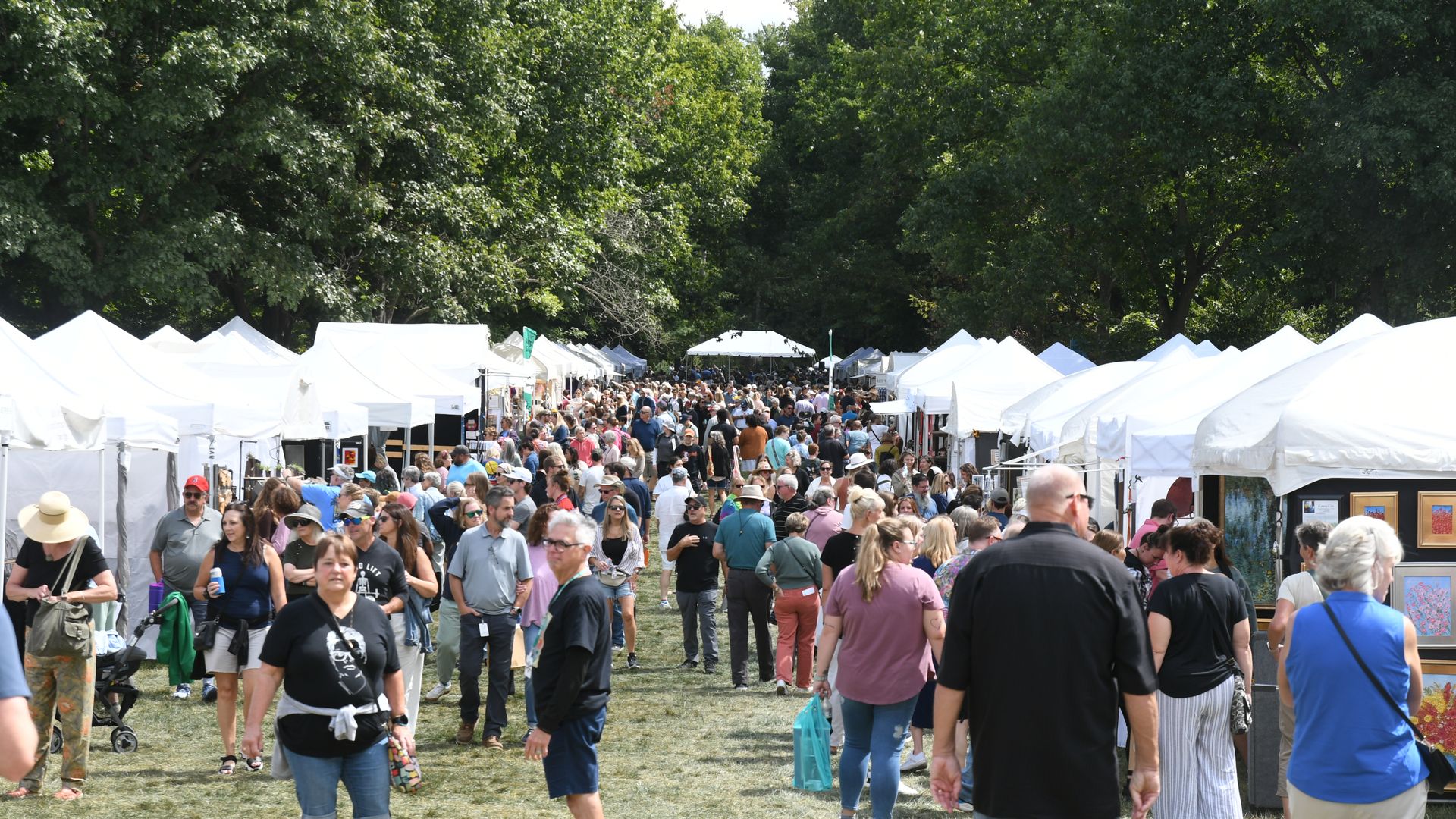 Busy outdoor market scene with white vendor tents lining both sides, a dense crowd walking along a central path under tall green trees on a sunny day.