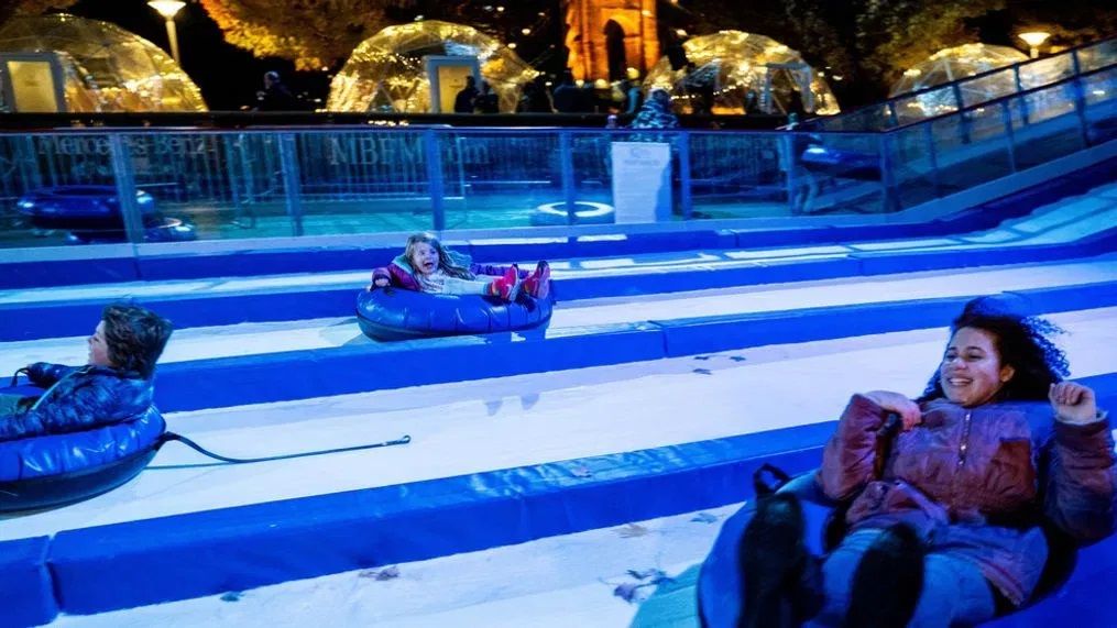 Three kids snow tubing against a lighted festival backdrop 