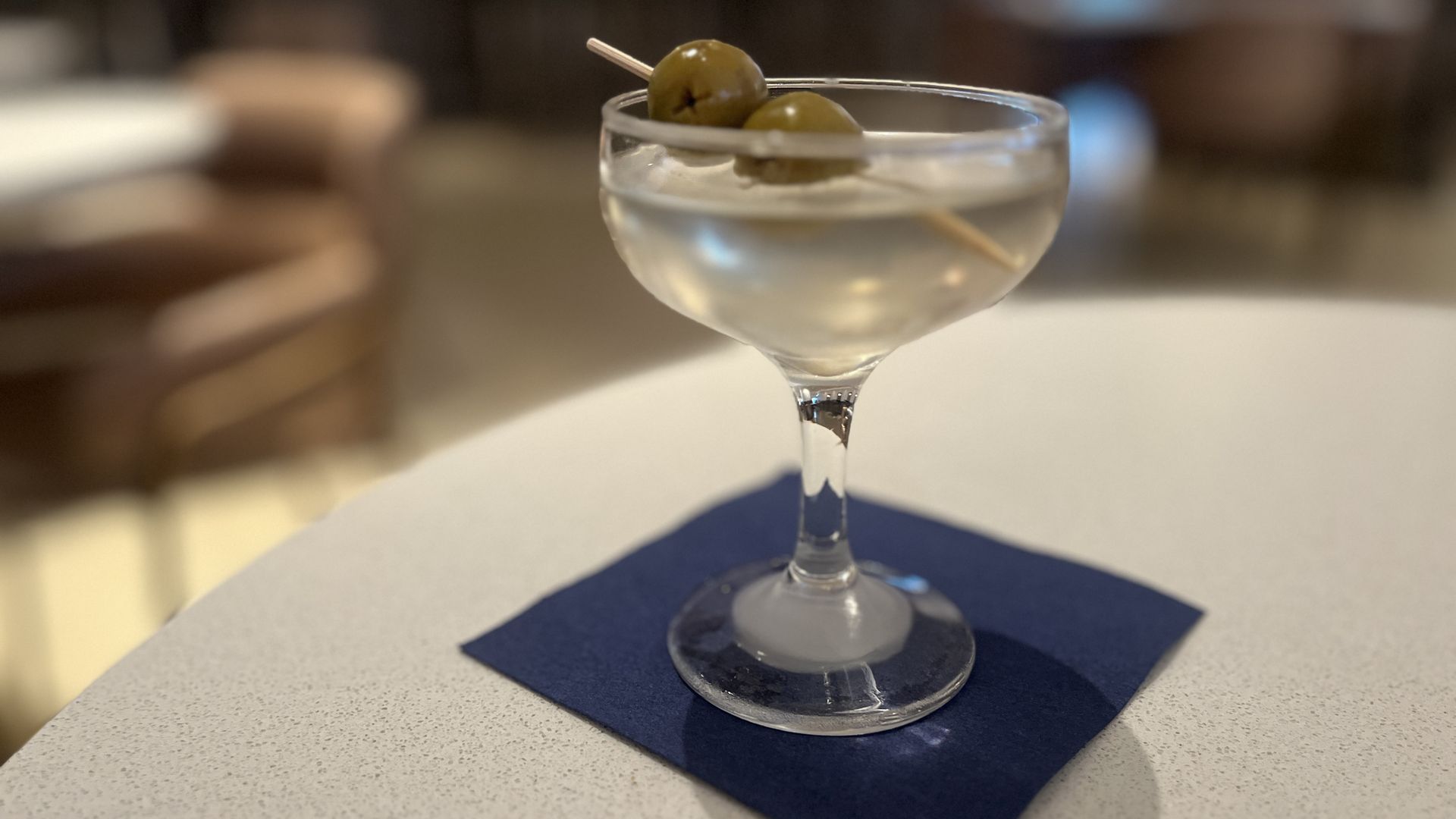 A clear martini in a coupe glass, garnished with two green olives on a toothpick, resting on a dark blue napkin on a speckled white table, with a blurred background.