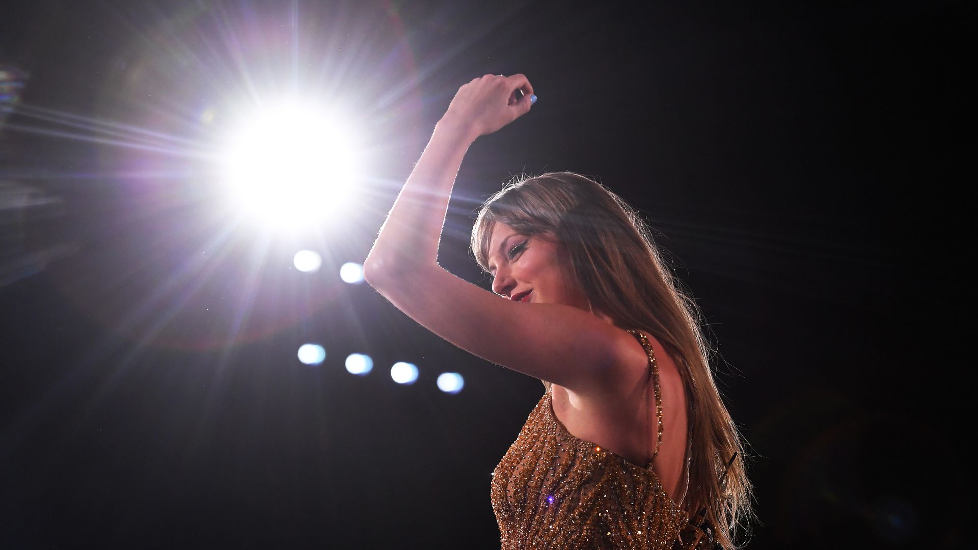 Taylor Swift smiles with her arm up in the air on stage. 
