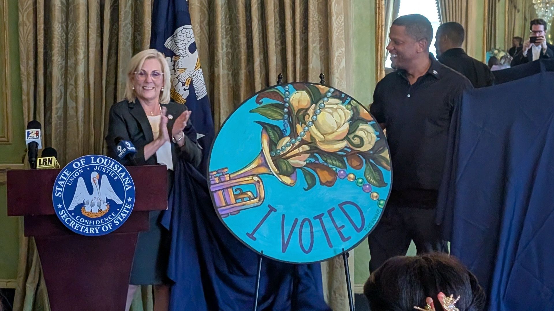 An ornate green and gold room with a Louisiana Secretary of State podium. A woman speaks as a man holds a large "I VOTED" sign; US flag left, reporters and a crystal chandelier in back.