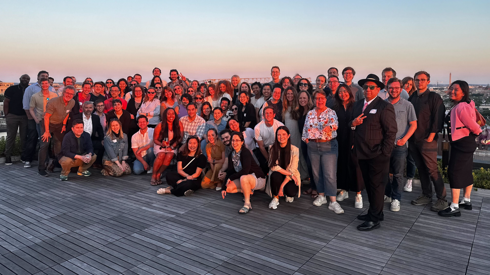 A larger group photo of many people, on an outdoor deck, with a city skyline in the background.