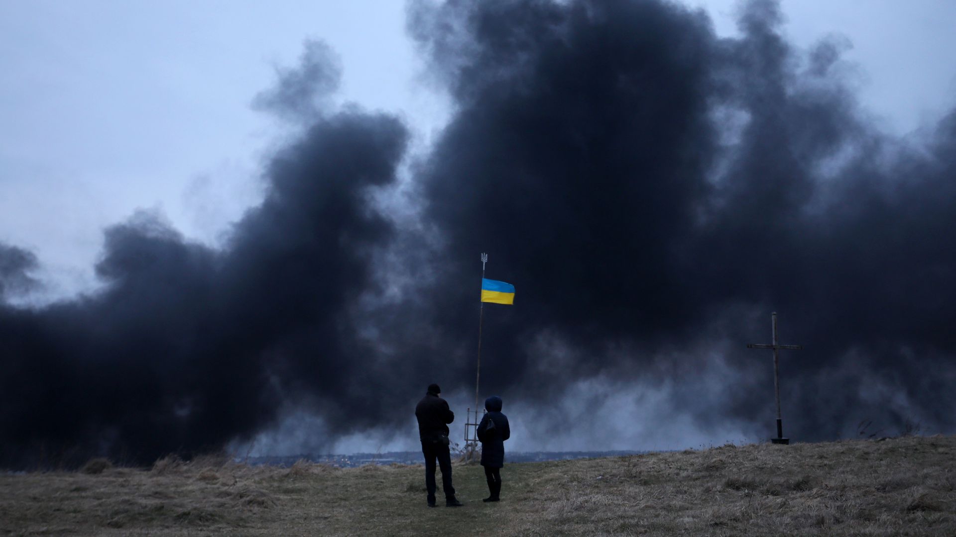 People stand in front of an Ukrainian national flag fluttering as dark smoke and flames rise from a fire following an air strike in the western Ukrainian city of Lviv, on March 26.