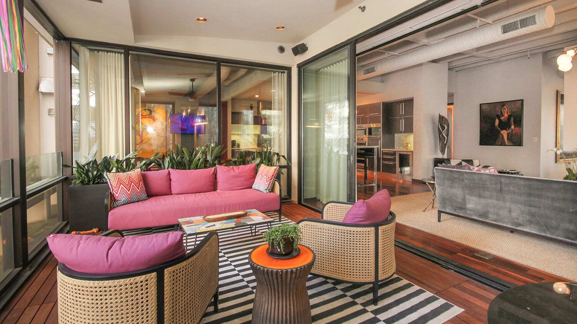 Porch converted into an outdoor living space. It has large floor-to-ceiling glass doors and the pink couches on a black and white striped rug. 