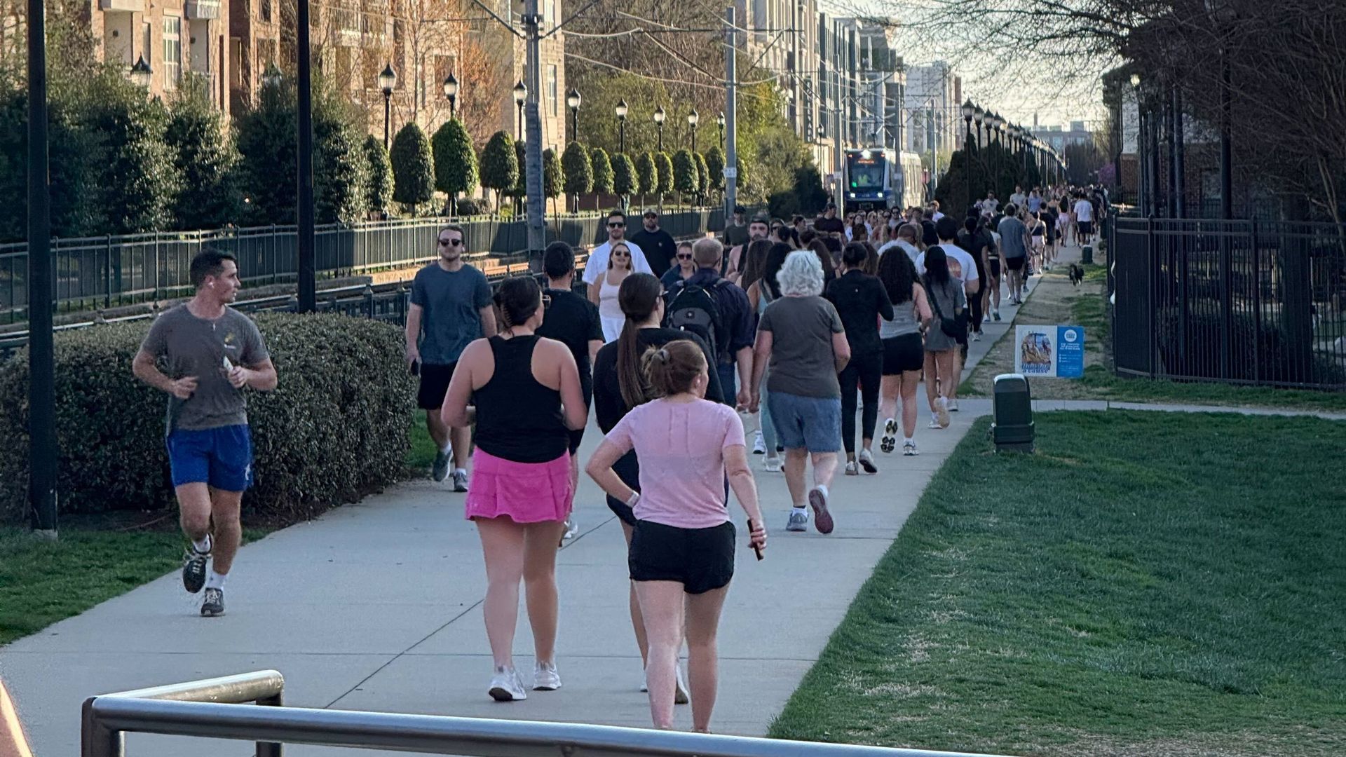 Sunlit city sidewalk flanked by beige apartment buildings on the left and bare trees on the right. A crowd of pedestrians and joggers fills the path, with shrubs and street lamps along the route.