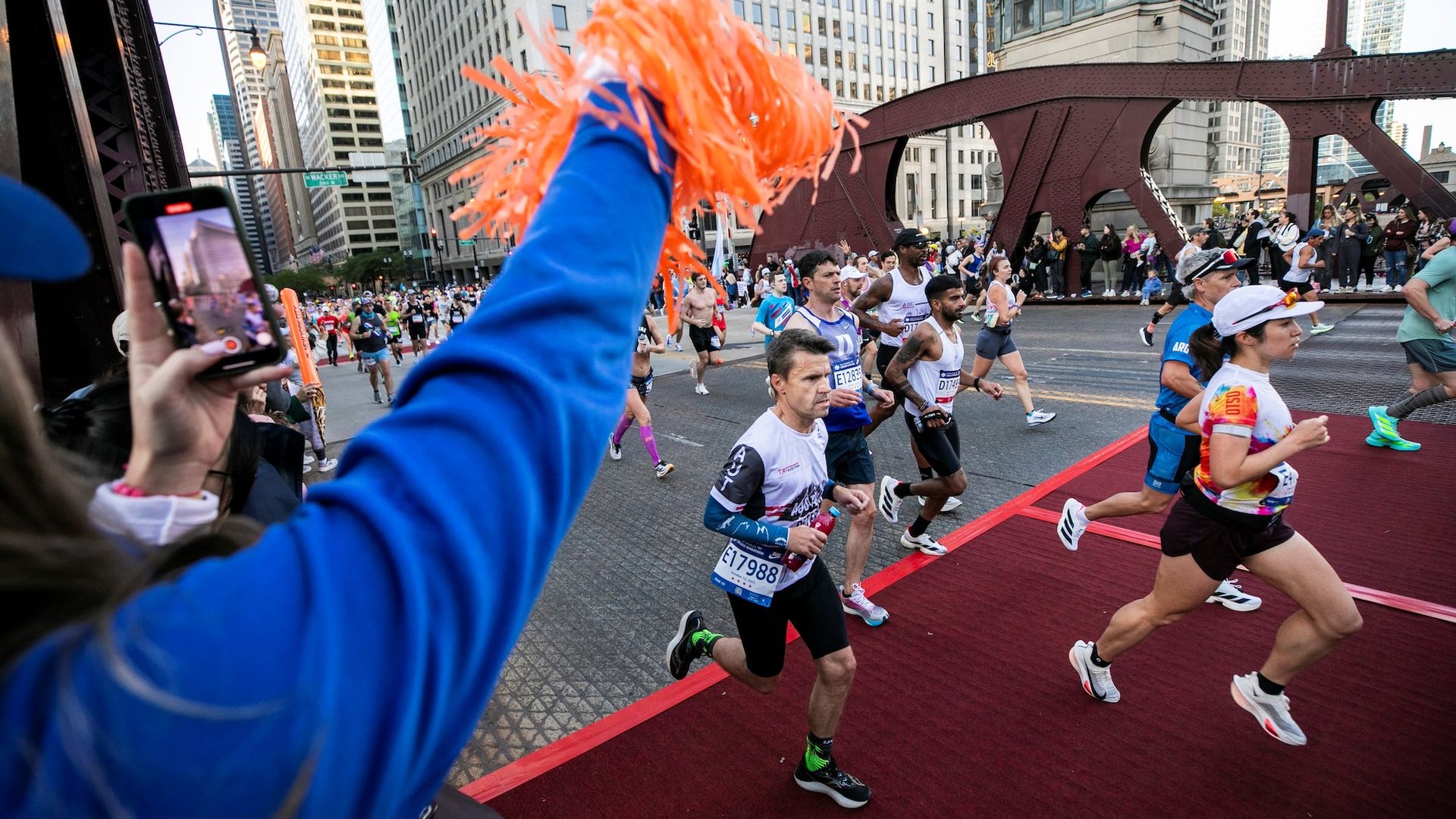 Runners in a city marathon pass under a steel bridge on a red-carpet finish; a spectator in a blue jacket raises orange pom-poms as crowds line the street.