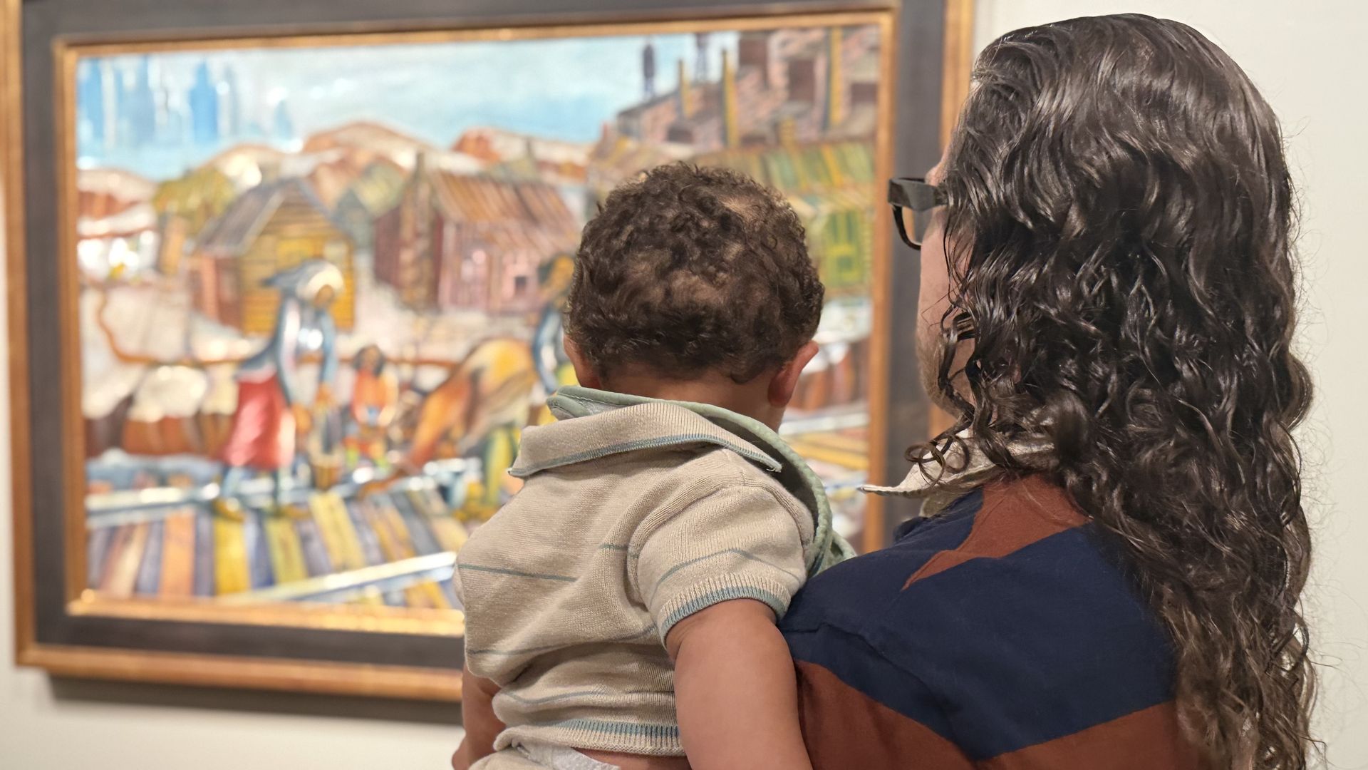 Back view of a man with long, curly hair and glasses holding a young child as they gaze at a colorful painting in a framed display on a gallery wall.
