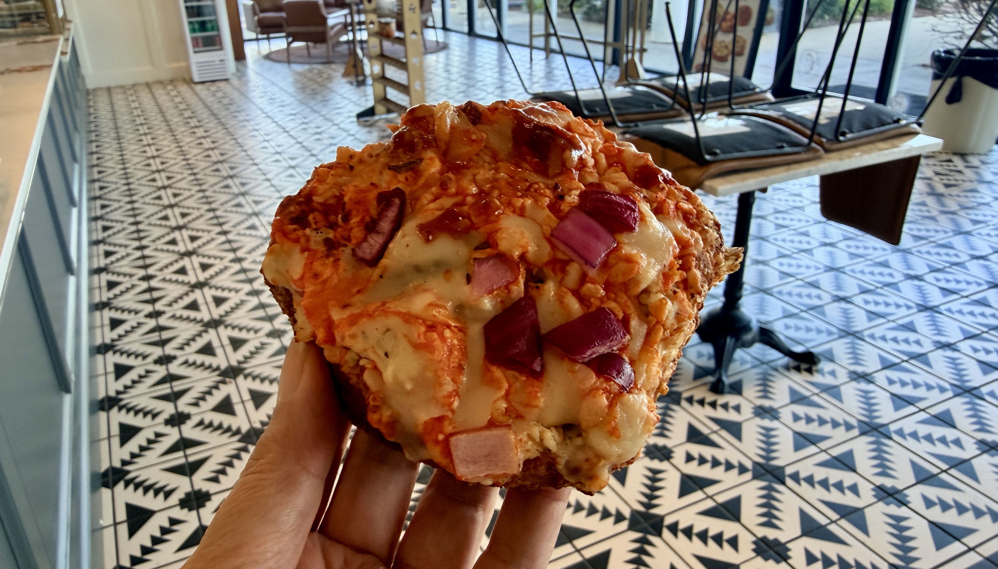 Close-up of a large crumb-coated savory pastry with orange crumbs and pink ham cubes, held in a hand inside a bright cafe with a geometric black-and-white tiled floor.